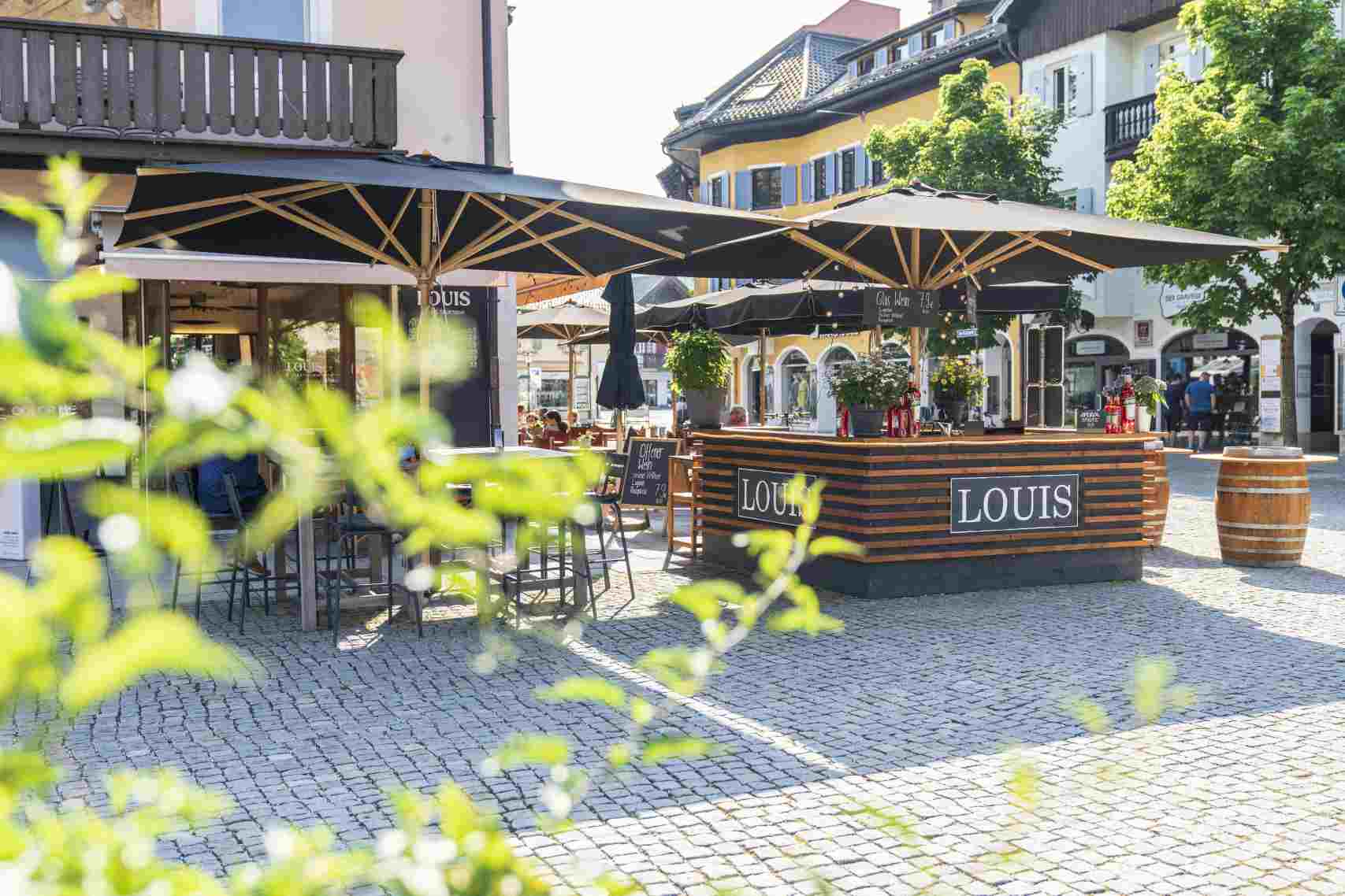 Weishäupl Klassiker Schirm: Außenbereich eines Cafés mit großen, schwarzen Sonnenschirmen und Holztischen auf sonnigem, gepflastertem Platz.