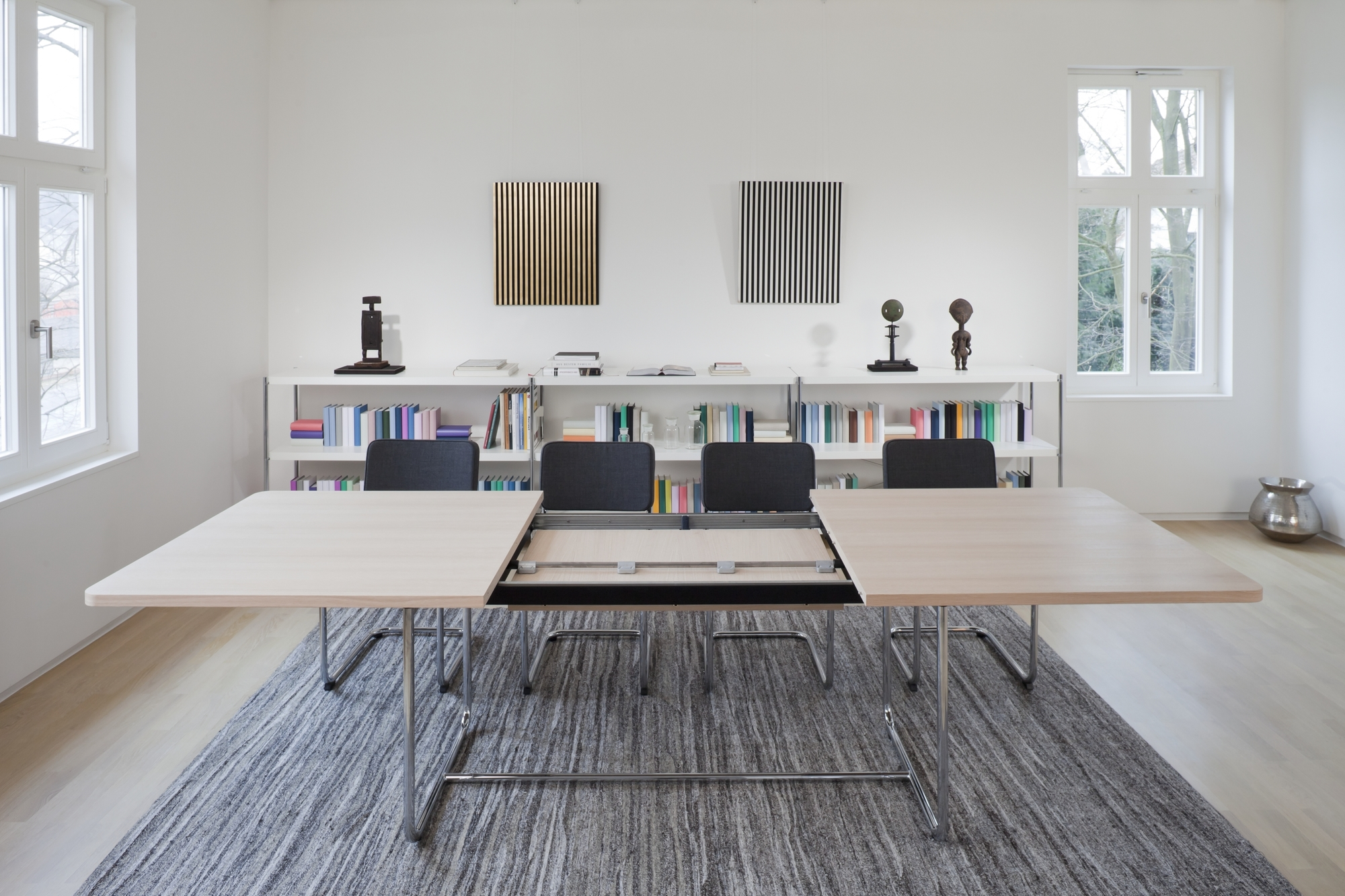 S 1072 Thonet Esstisch ausziehbar, helle Tischplatte, Chromgestell, teilweise ausgezogen, mit Stühlen und Bücherregal im Hintergrund.