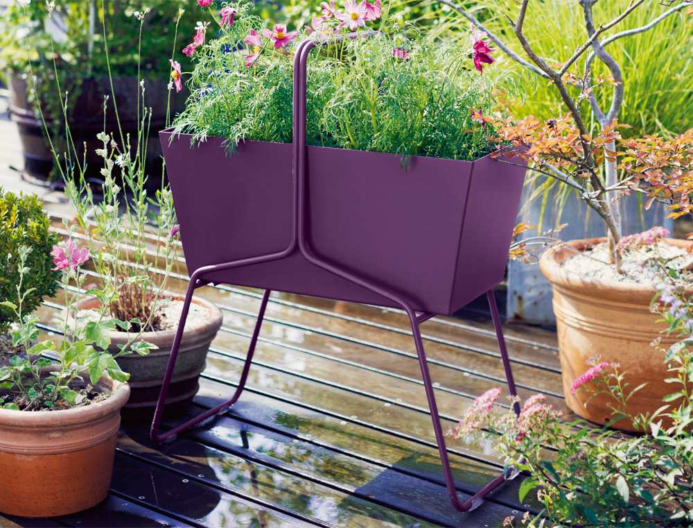 Fermob Basket Blumenkasten hoch, lila, mit Blumen bepflanzt, auf einer Holzterrasse, Gartendekoration.