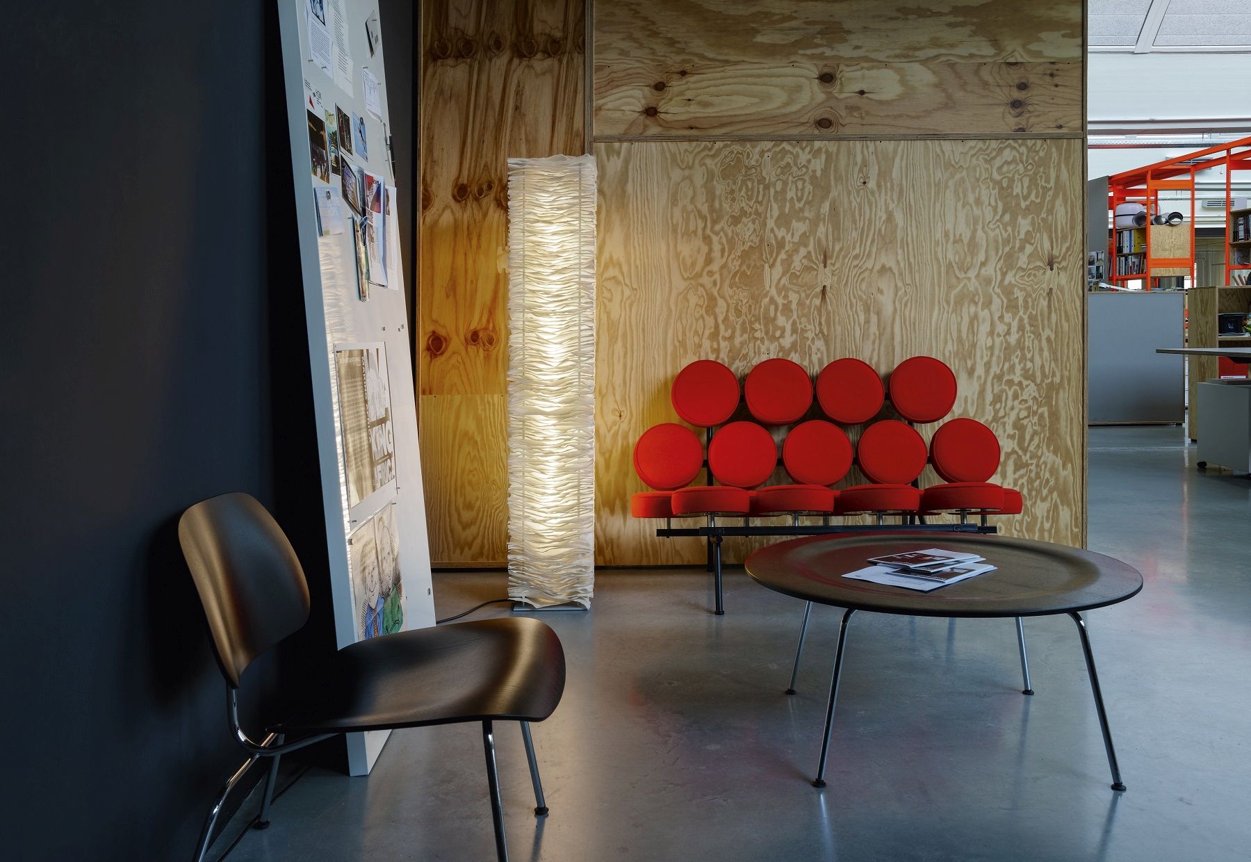 Moderner Büroraum mit rotem Sofa, Design-Stehlampe, braunem Stuhl und rundem Couchtisch auf Betonboden.