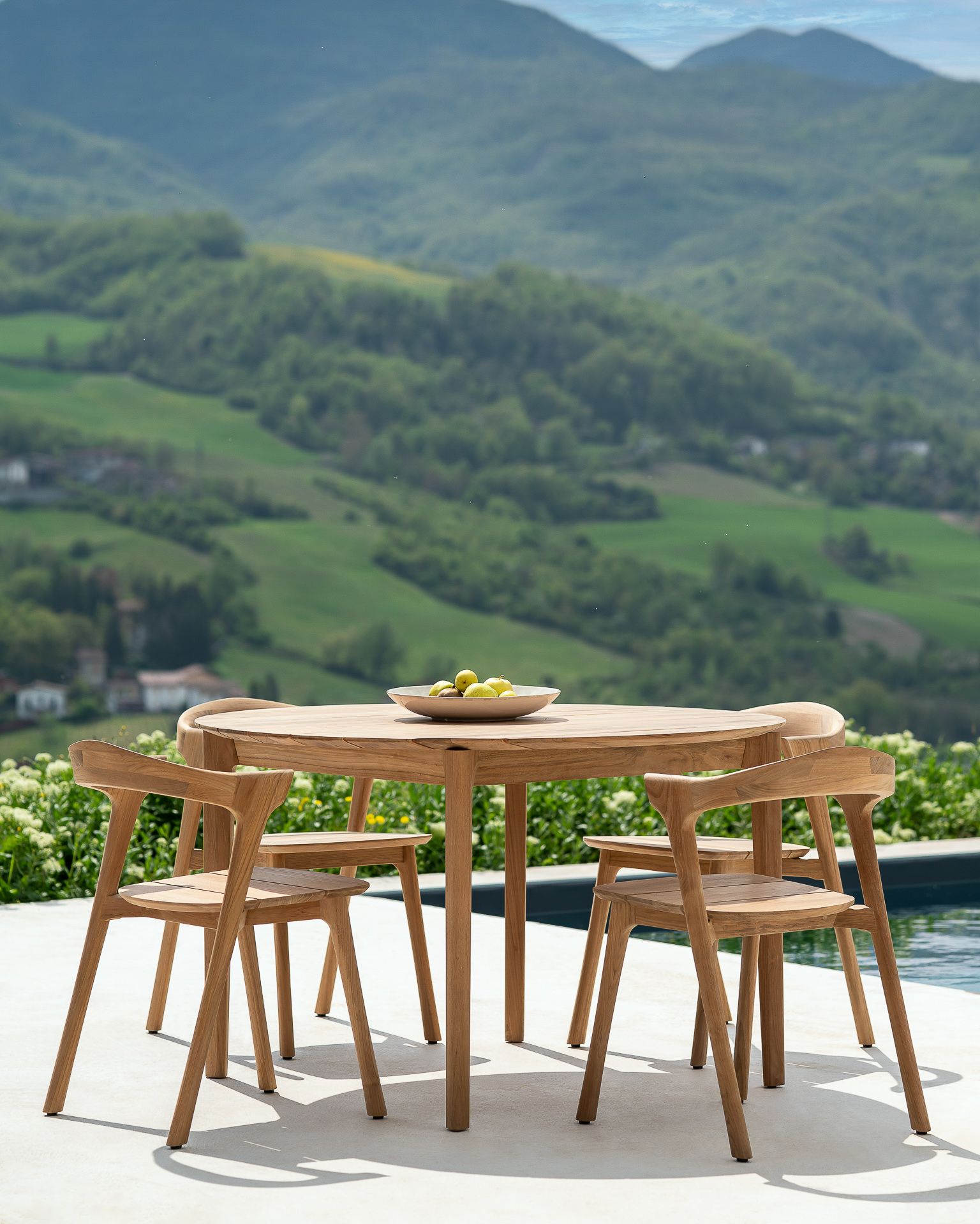 Ethnicraft Bok Teak Gartentisch mit vier Stühlen vor malerischer Hügellandschaft für entspannte Stunden im Freien.