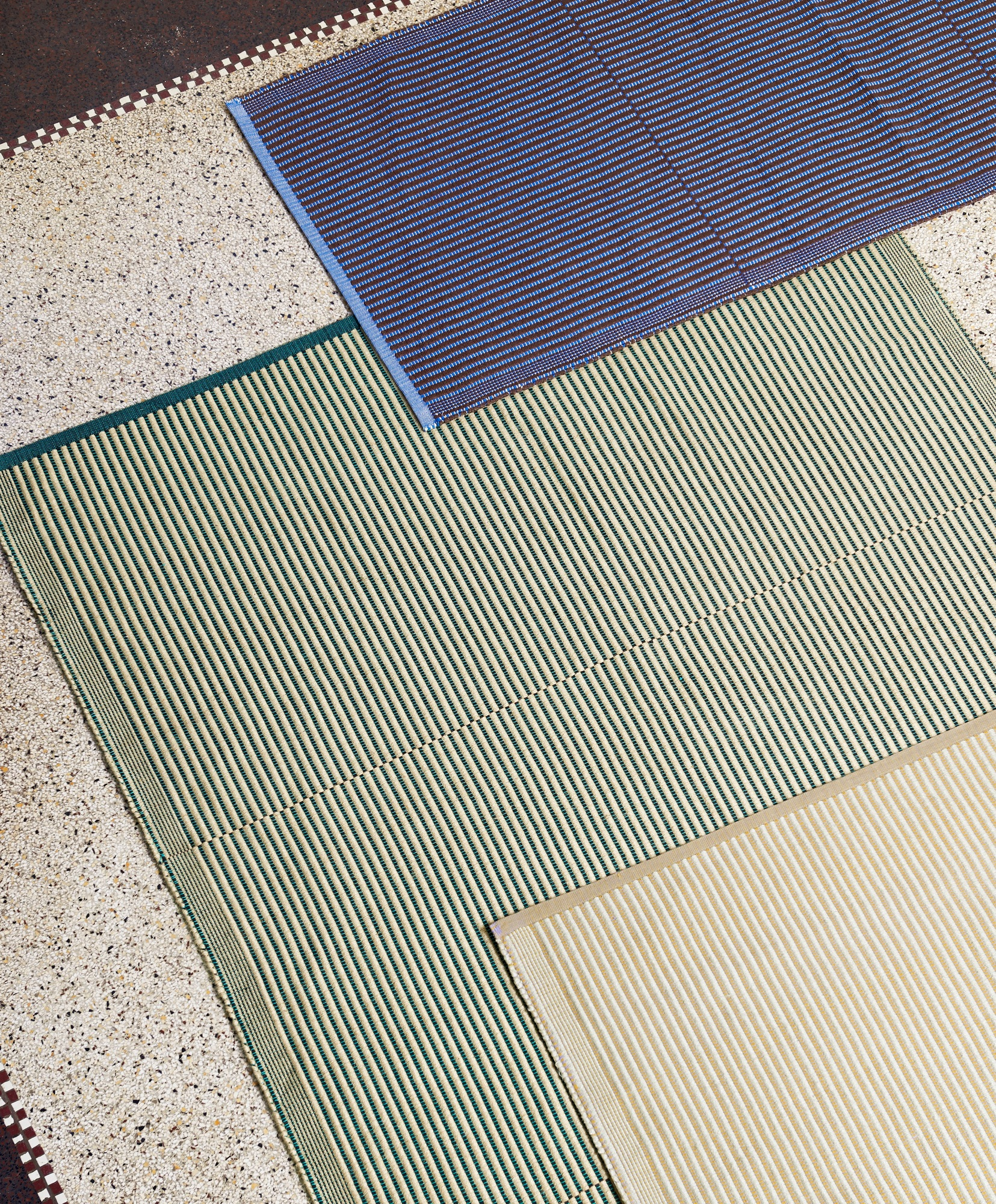 Nahaufnahme von drei gestreiften Hay Teppichen in Blau, Grün und Beige auf einem Terrazzoboden.