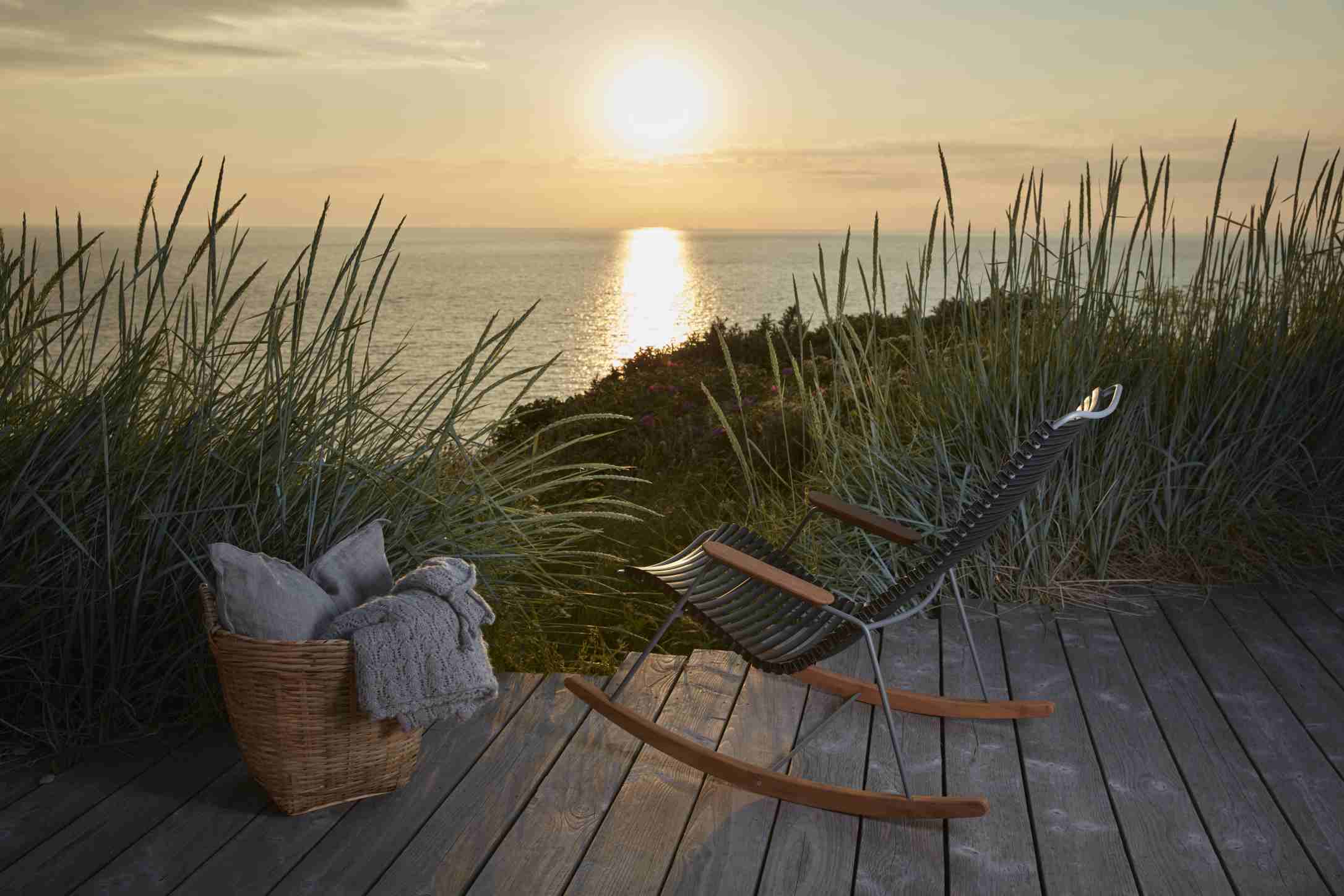 Houe Click Rocking Chair Outdoor Schaukelstuhl auf Holzterrasse mit Meerblick im Sonnenuntergang. Korb mit Kissen.