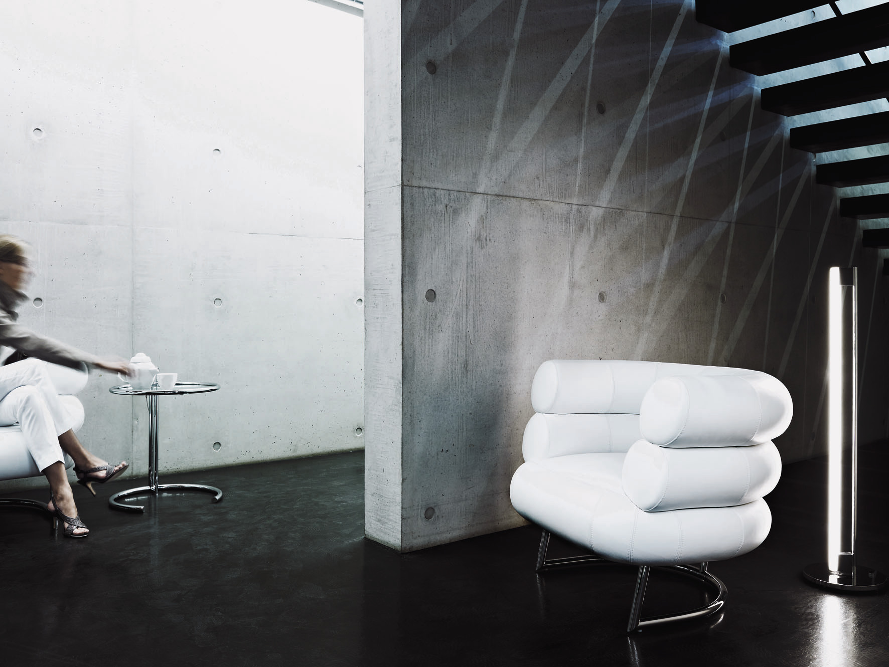 Moderner Wohnraum mit Adjustable Table E1027 Beistelltisch, Designklassiker mit Glasplatte und Stahlrohrgestell neben weißem Sessel.