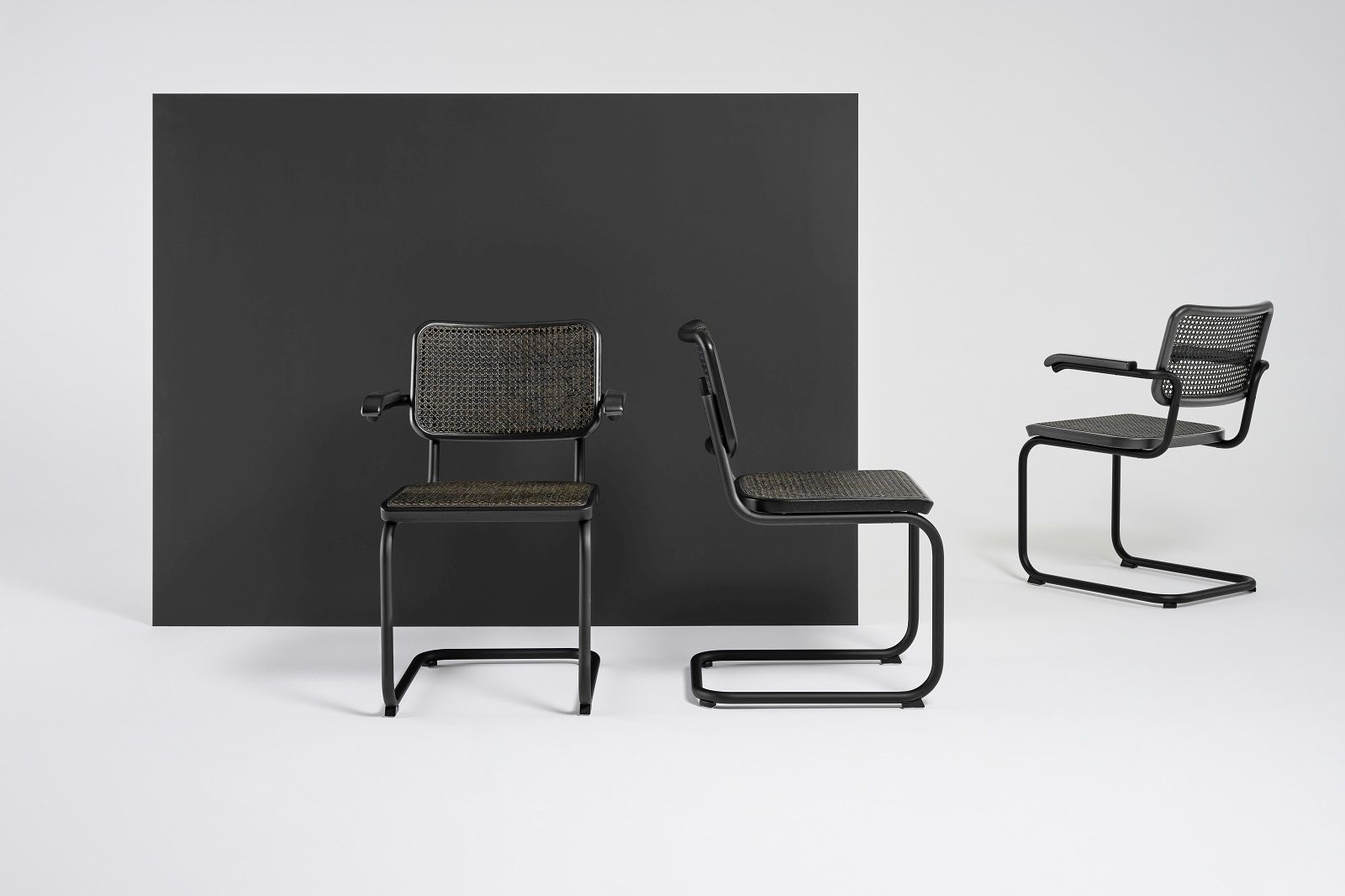 Zwei schwarze Thonet S 32 Freischwinger Stühle mit Armlehnen und Wiener Geflecht vor schwarzem Hintergrund.