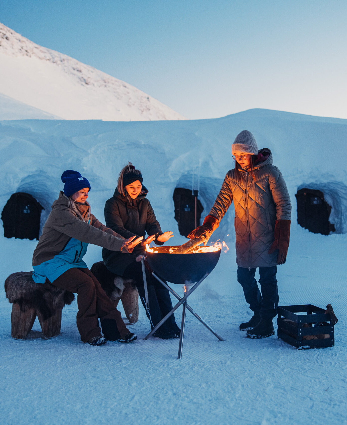 Drei Personen wärmen sich an einer Feuerschale im Schnee vor Iglus. Winterurlaub und Outdoor-Aktivitäten.