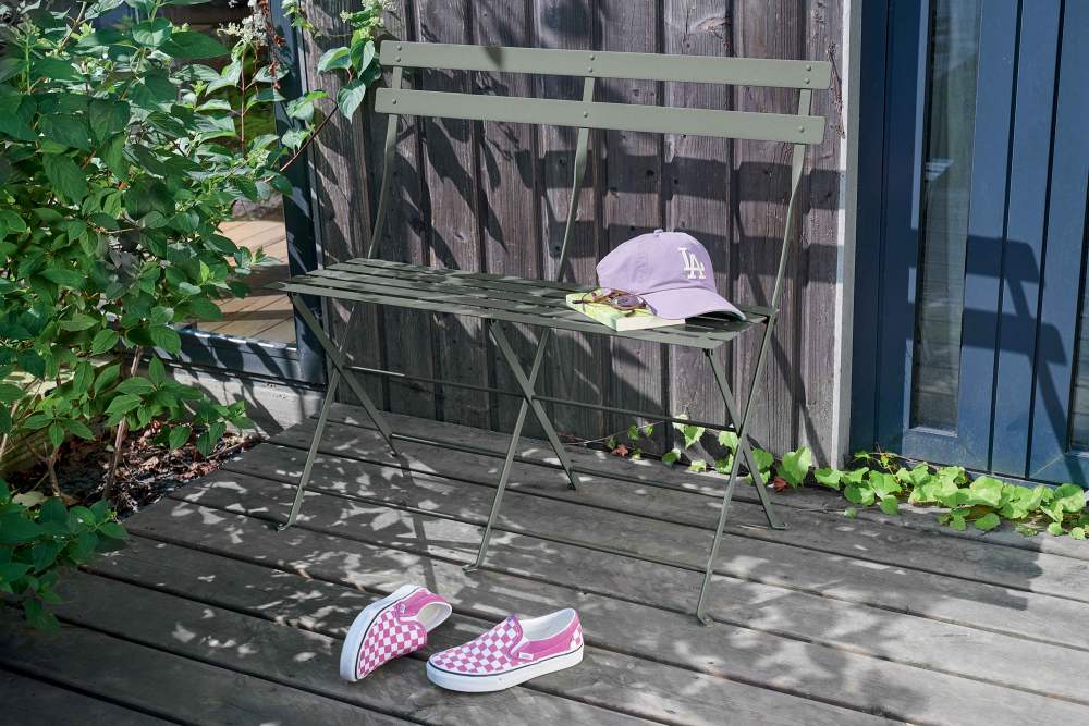 Grüne Fermob Bistro Klappbank für zwei Personen im Außenbereich mit Accessoires auf Holzterrasse.