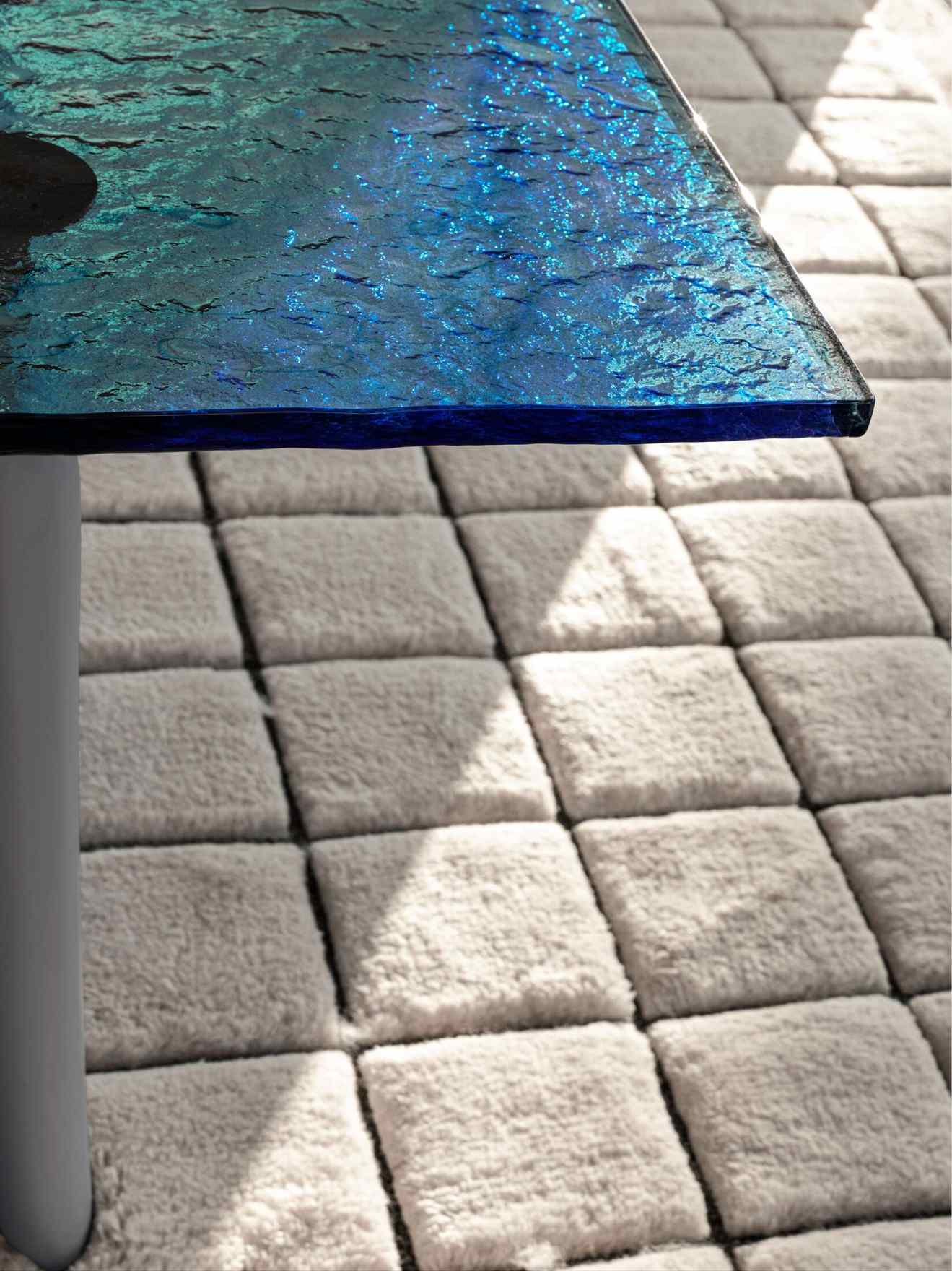 Detailaufnahme des Cassina LC6 Esstisches mit blauer Glasplatte und grauem Teppich im Hintergrund.