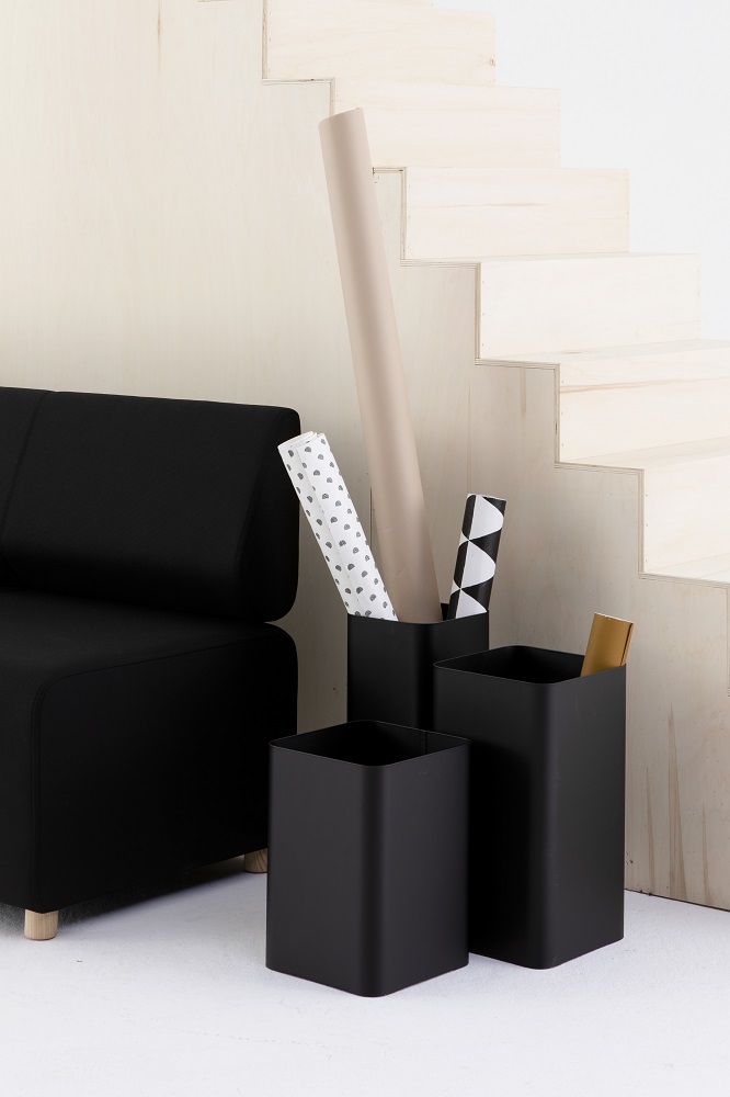 Drei schwarze Cascando Flow Papierkörbe unterschiedlicher Größe neben einem schwarzen Sofa und einer Holztreppe.