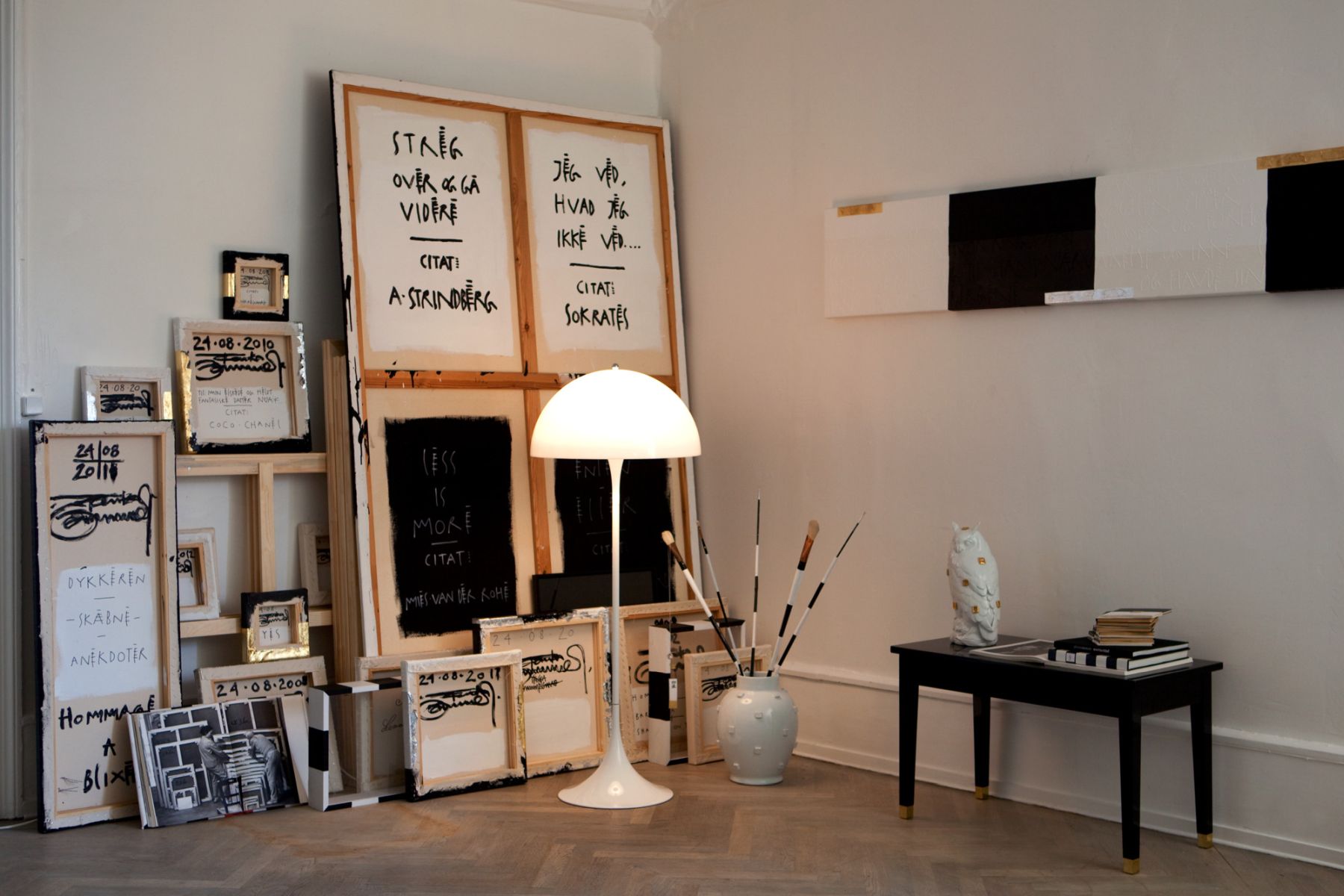 Innenraum mit Kunstwerken, Staffeleien, weißer Stehlampe und schwarzem Tisch mit Dekoration.