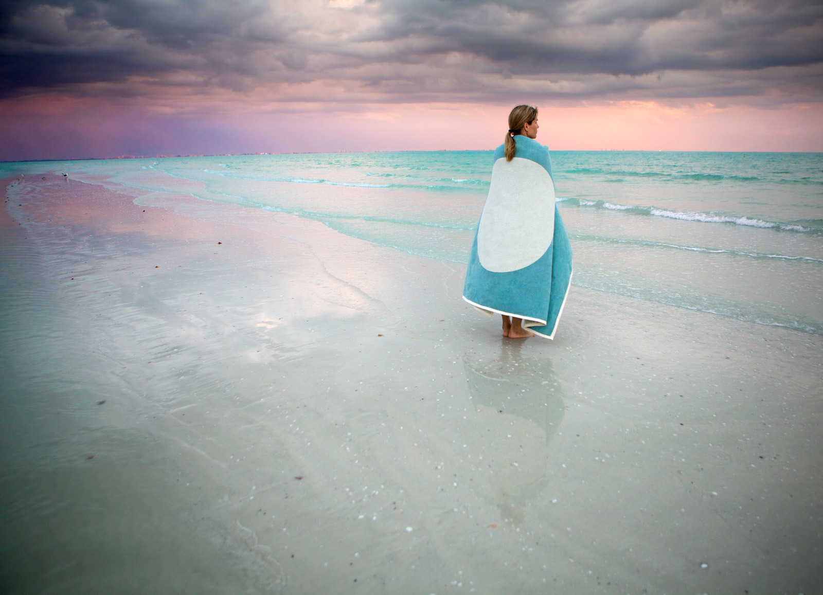 Frau am Strand in die blaue Vera Wolldecke von Pappelina gehüllt, hochwertige Wohndecke aus Schweden.