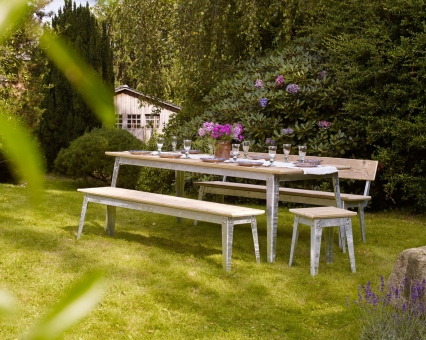 Gartentisch mit Bänken und Hocker aus hellem Holz auf Rasen, gedeckt mit Blumen und Gläsern.