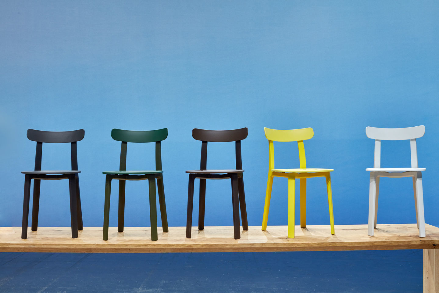 Fünf Vitra All Plastic Chairs Outdoor in verschiedenen Farben auf einem Holzbalken vor blauem Hintergrund.