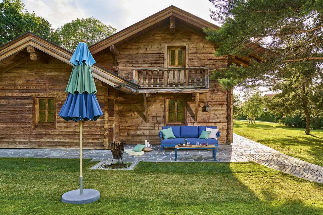 Blau-grüner Weishäupl Schirm vor einem Holzhaus. Klassischer Sonnenschirm für Garten und Terrasse.