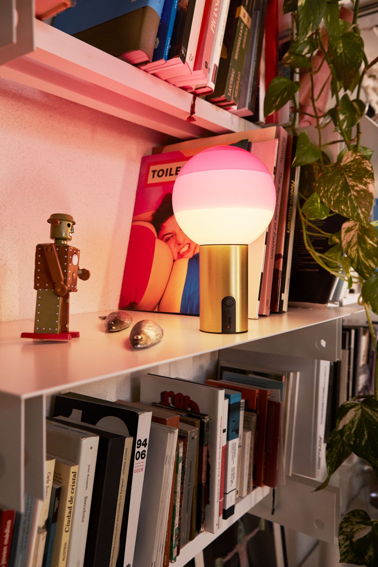 Goldene Tischleuchte mit rosa-weißem Lampenschirm auf einem Bücherregal, dekorative Beleuchtung für Wohnzimmer und Schlafzimmer.