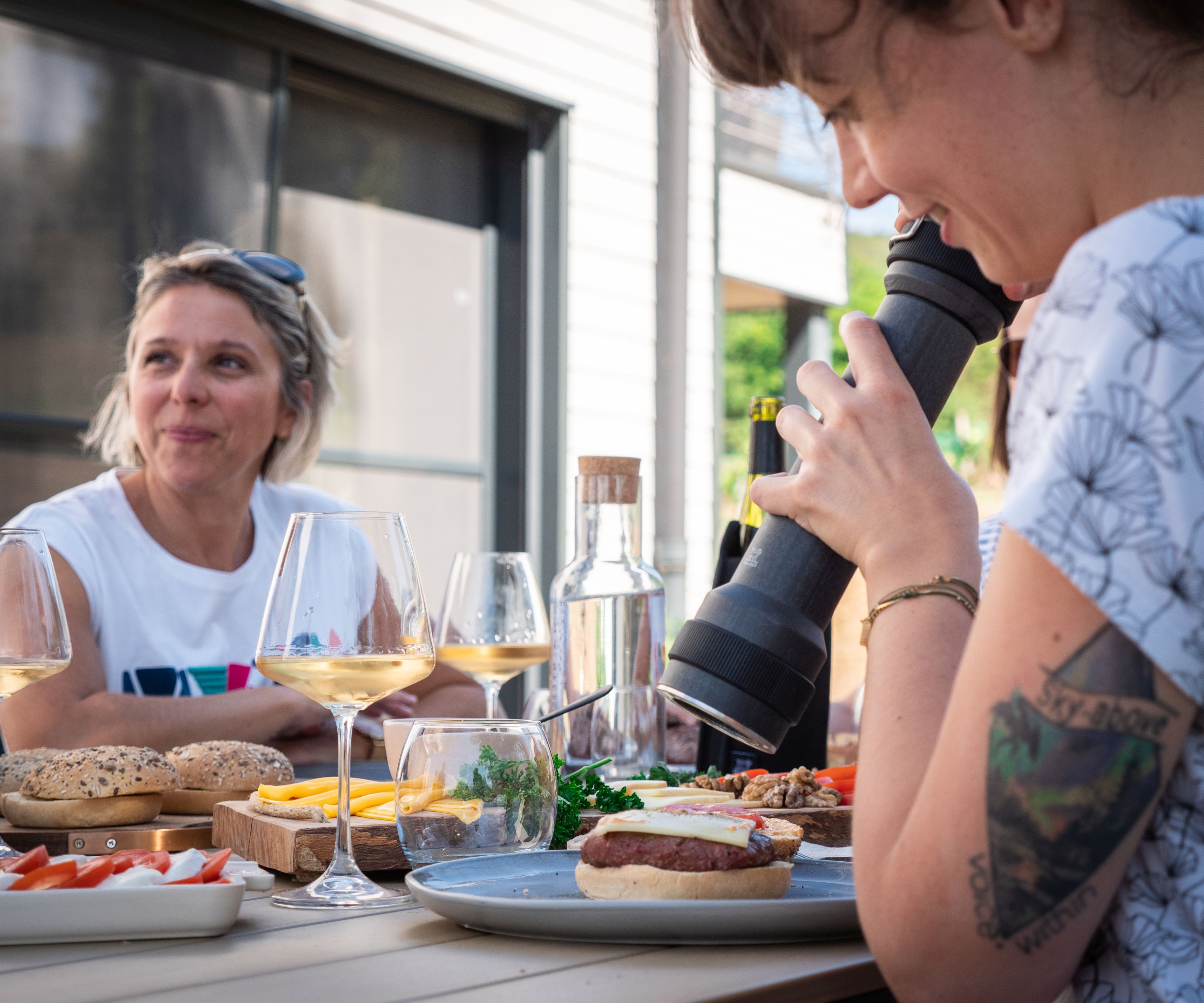 Frau würzt Burger mit Peugeot BBQ Pfeffermühle, Outdoor-Szene mit Freunden, Grillen und Wein.