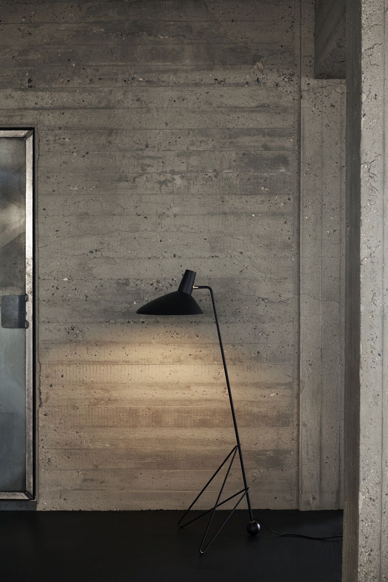 AndTradition Tripod HM8 Stehleuchte in Schwarz vor einer Betonwand, minimalistisches Design.