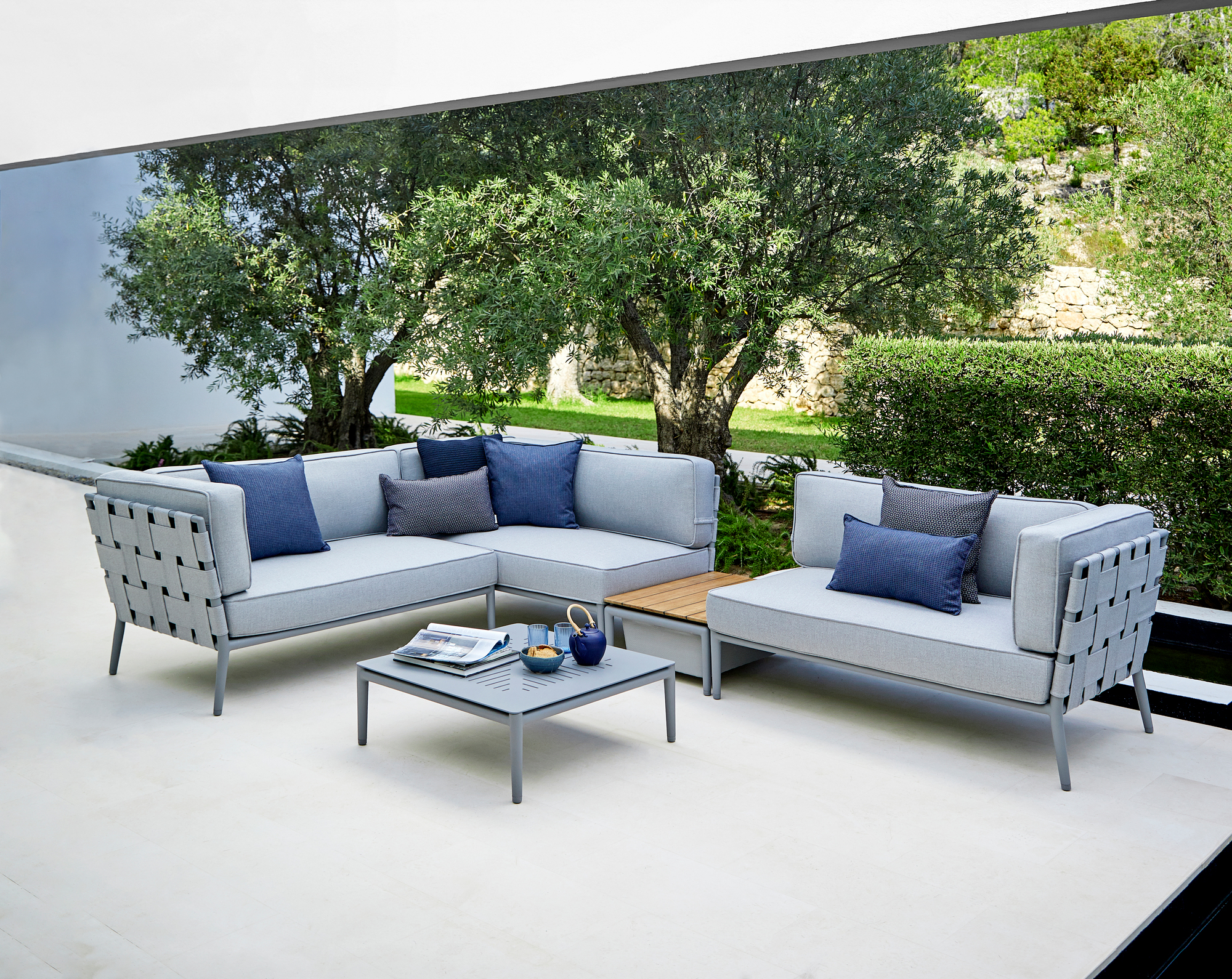 Conic AirTouch Outdoor Sofa, hellgraues 2-Sitzer Modulsofa rechts, mit blauen Kissen auf einer Terrasse.