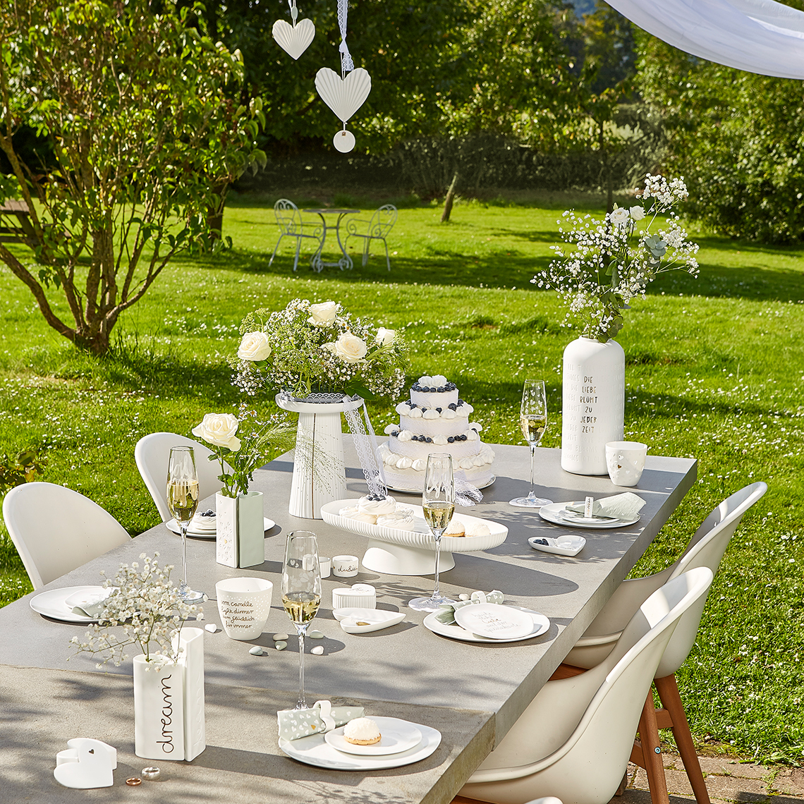 Hochzeitstafel im Garten mit "Die Liebe Blüht zu Jeder Zeit" Vase und Blumendeko für festliche Momente.