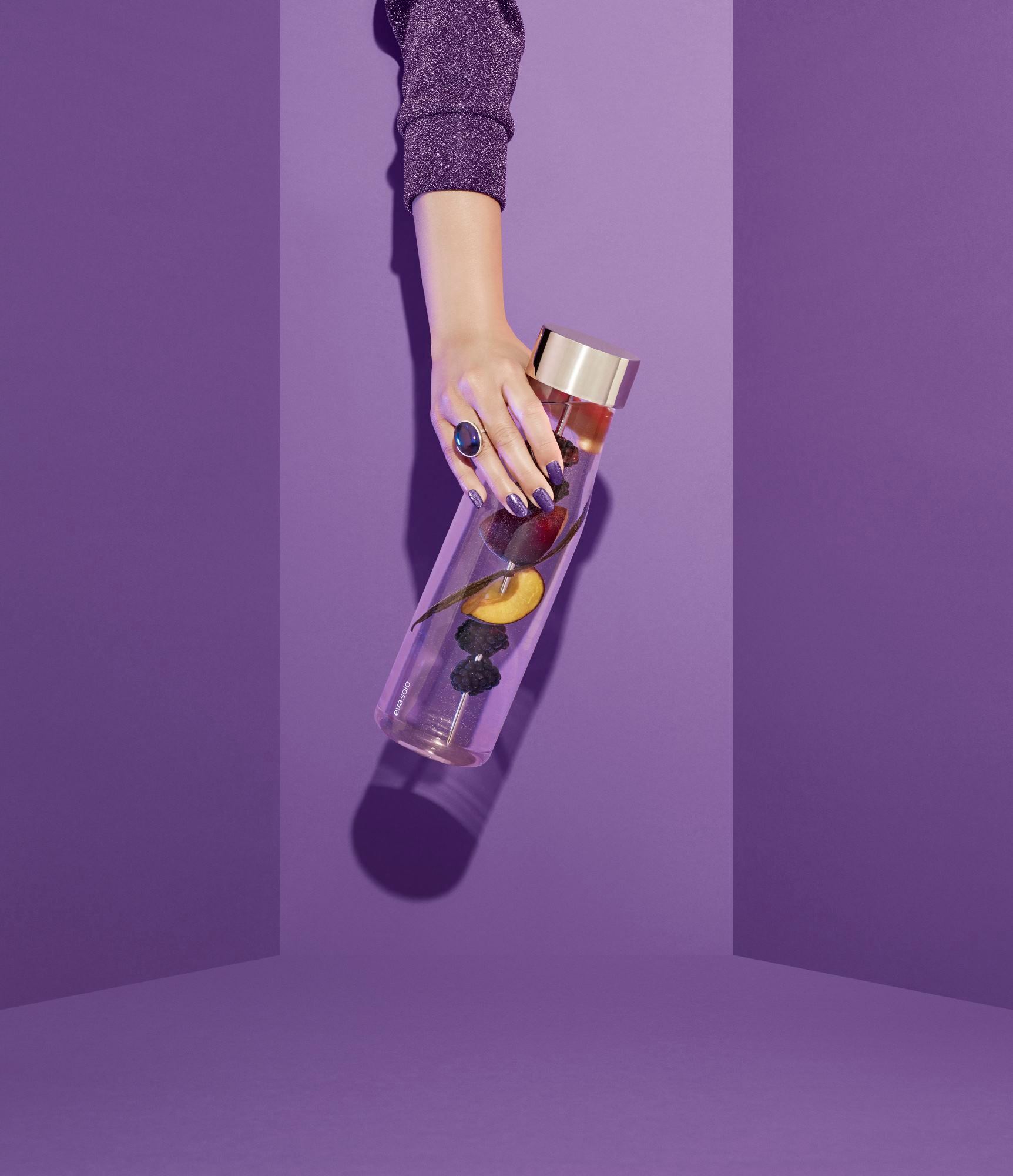 Hand hält My Flavour Karaffe von Eva Solo, gefüllt mit Früchten, vor lila Hintergrund.