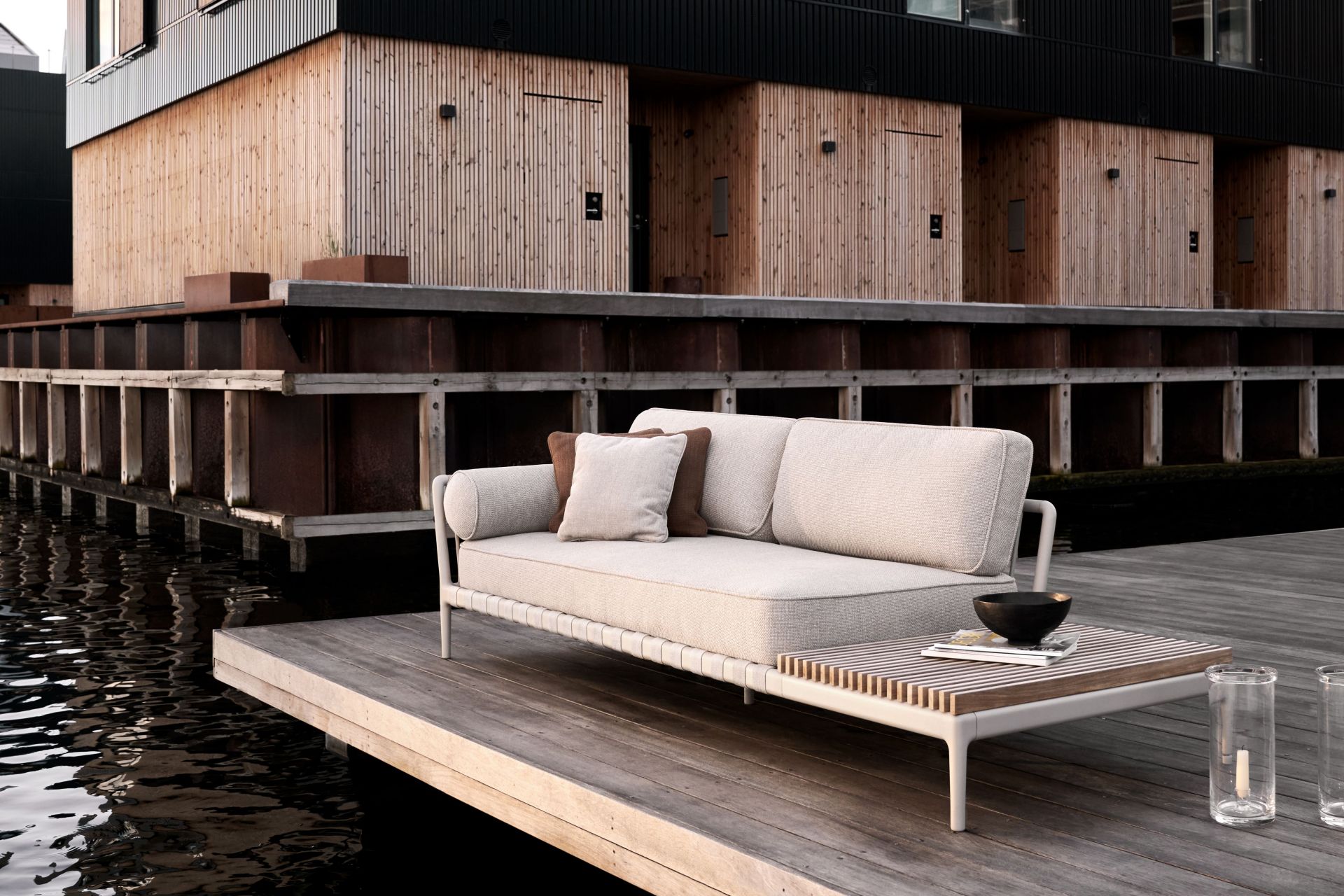 Beige Outdoor Sofa mit Kissen und Beistelltisch auf Holzterrasse am Wasser, modernes Gartenmöbel Set.
