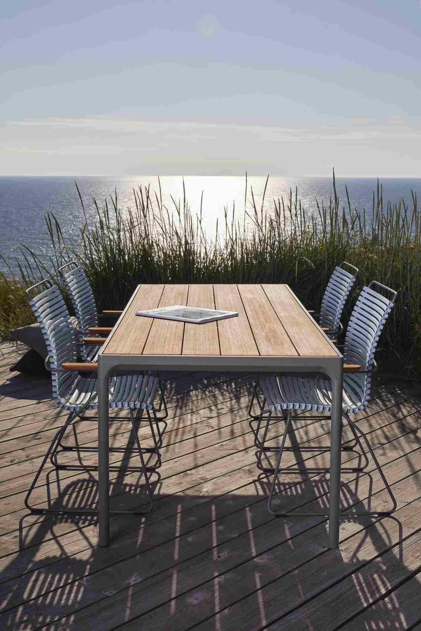Dunkelgraue Click Dining Stühle mit Armlehne von Houe und Holztisch auf einer Terrasse mit Meerblick.