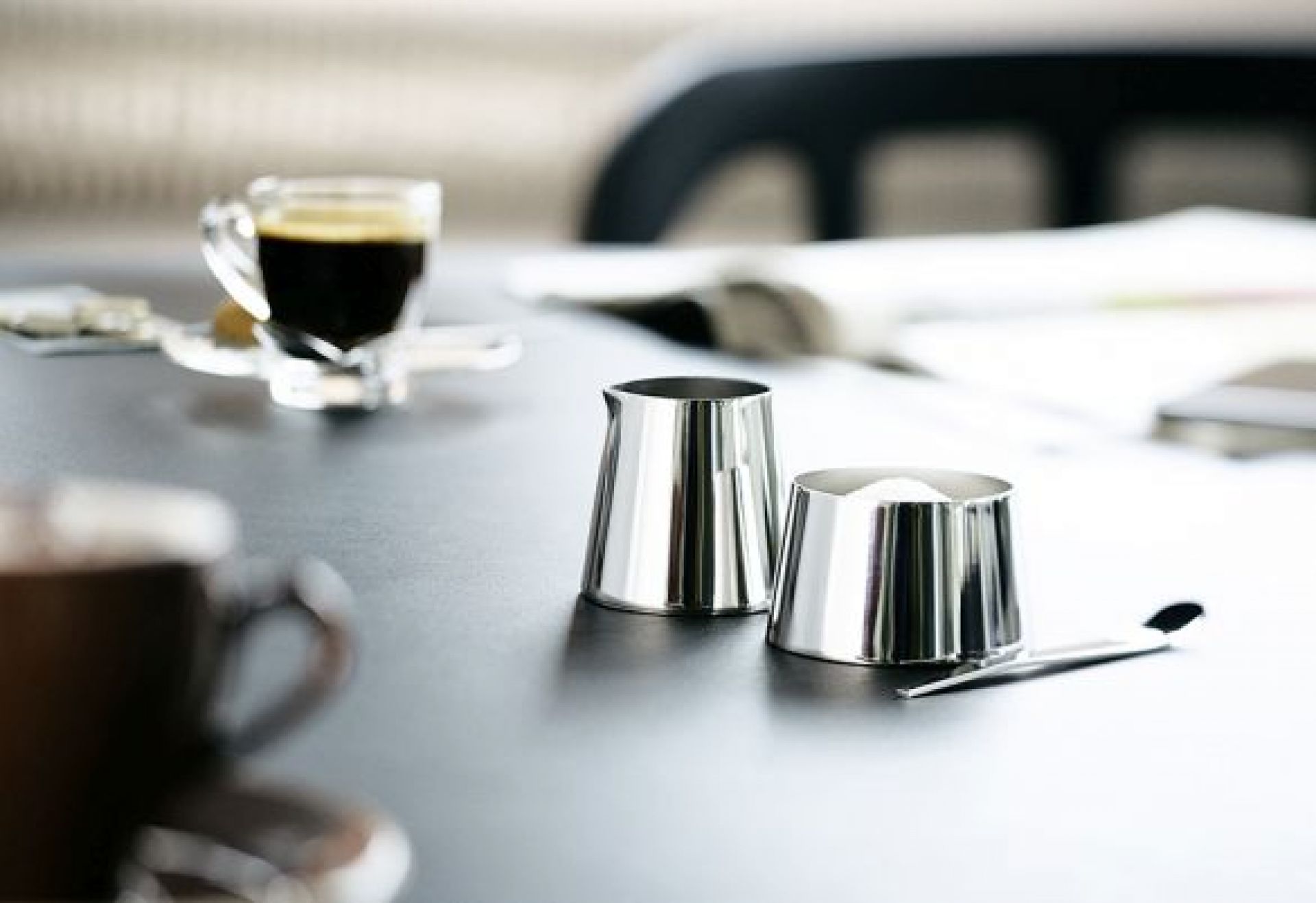 Nahaufnahme: Edelstahl Milchkanne und Zuckerdose mit Löffel, Kaffeetasse im Hintergrund.