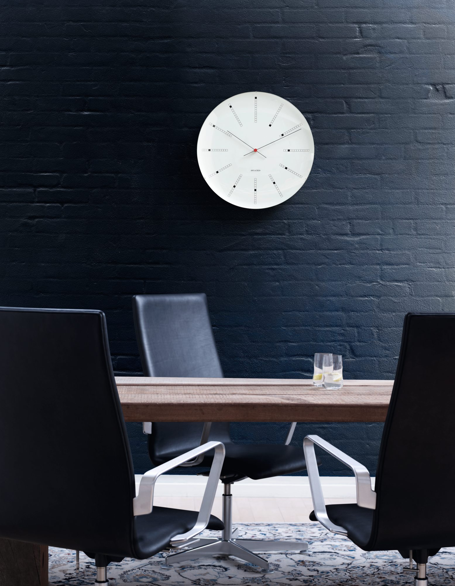 Bankers Wanduhr von Rosendahl Timepieces an dunkler Wand über Esstisch mit Stühlen.