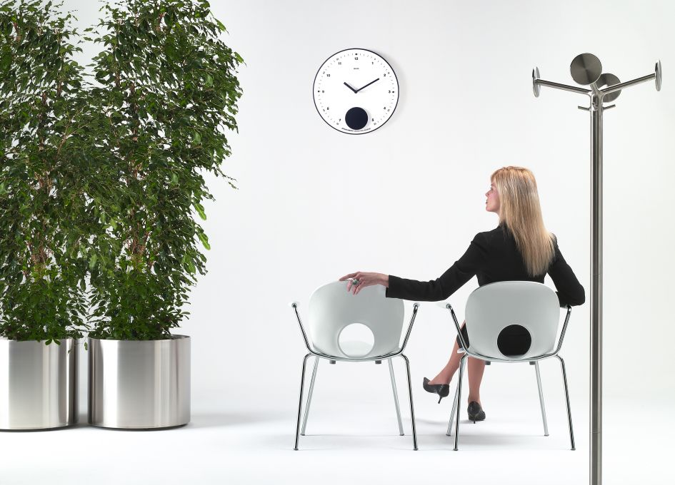 Moderne Rexite Wanduhr Appuntamento in einem minimalistischen Büro mit Frau, Pflanzen und Kleiderständer.