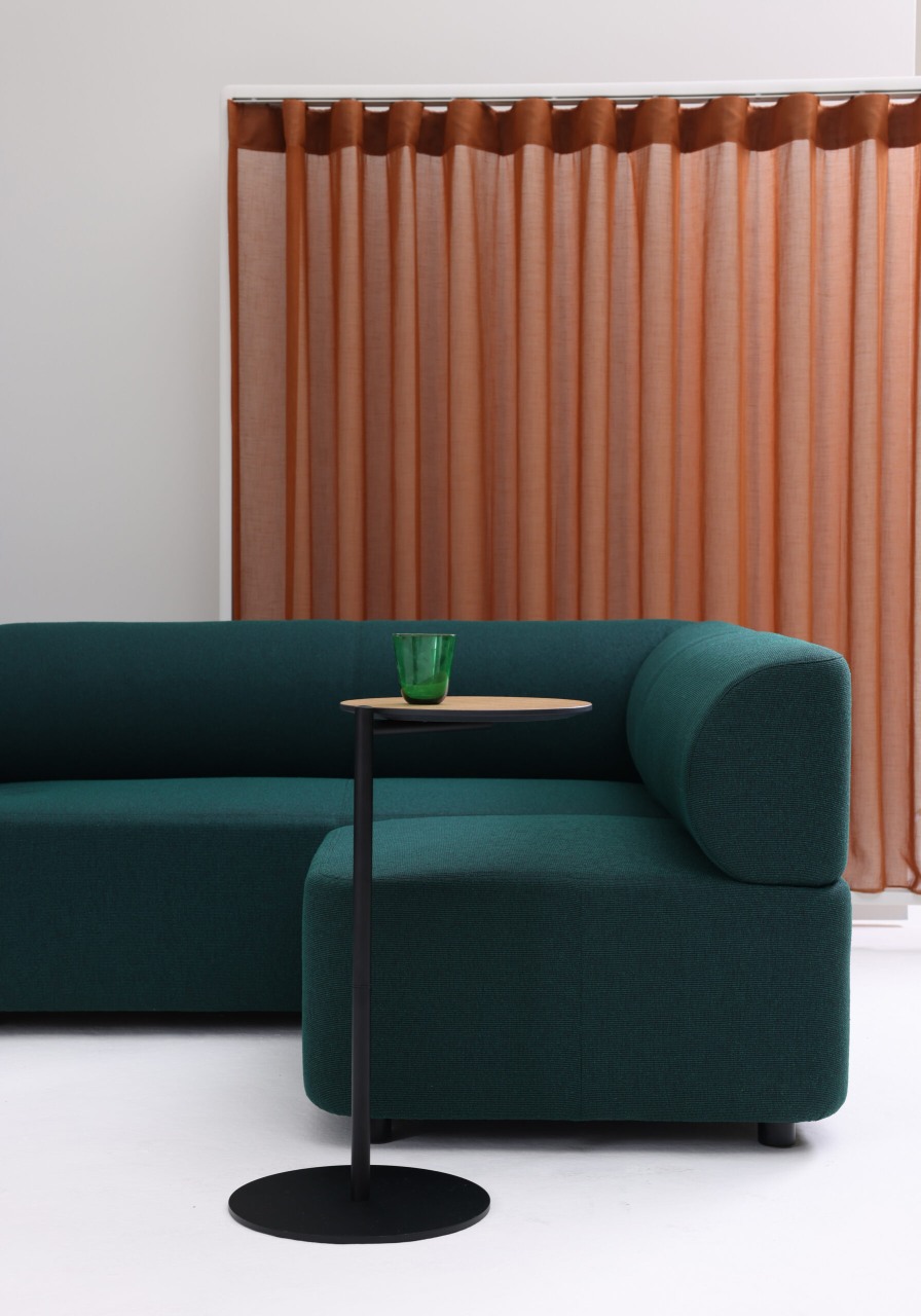 Grünes Cascando Mezza Sitzelement Sofa mit Beistelltisch und grünem Glas vor braunem Vorhang.