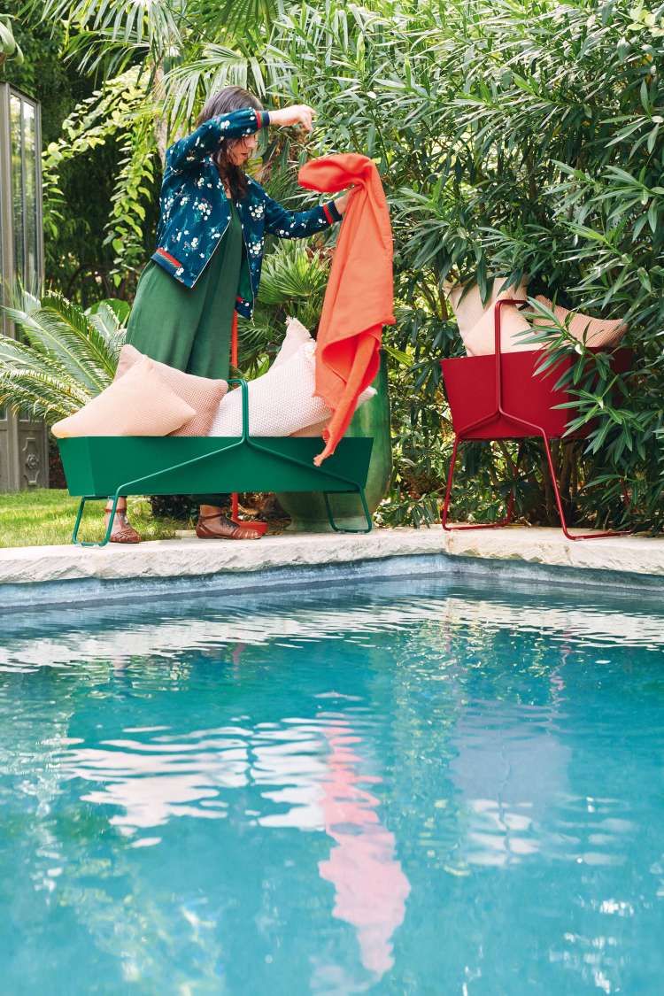 Frau drapiert Tuch über grünen Fermob Basket Blumenkasten hoch am Pool, roter Blumenkasten im Hintergrund.