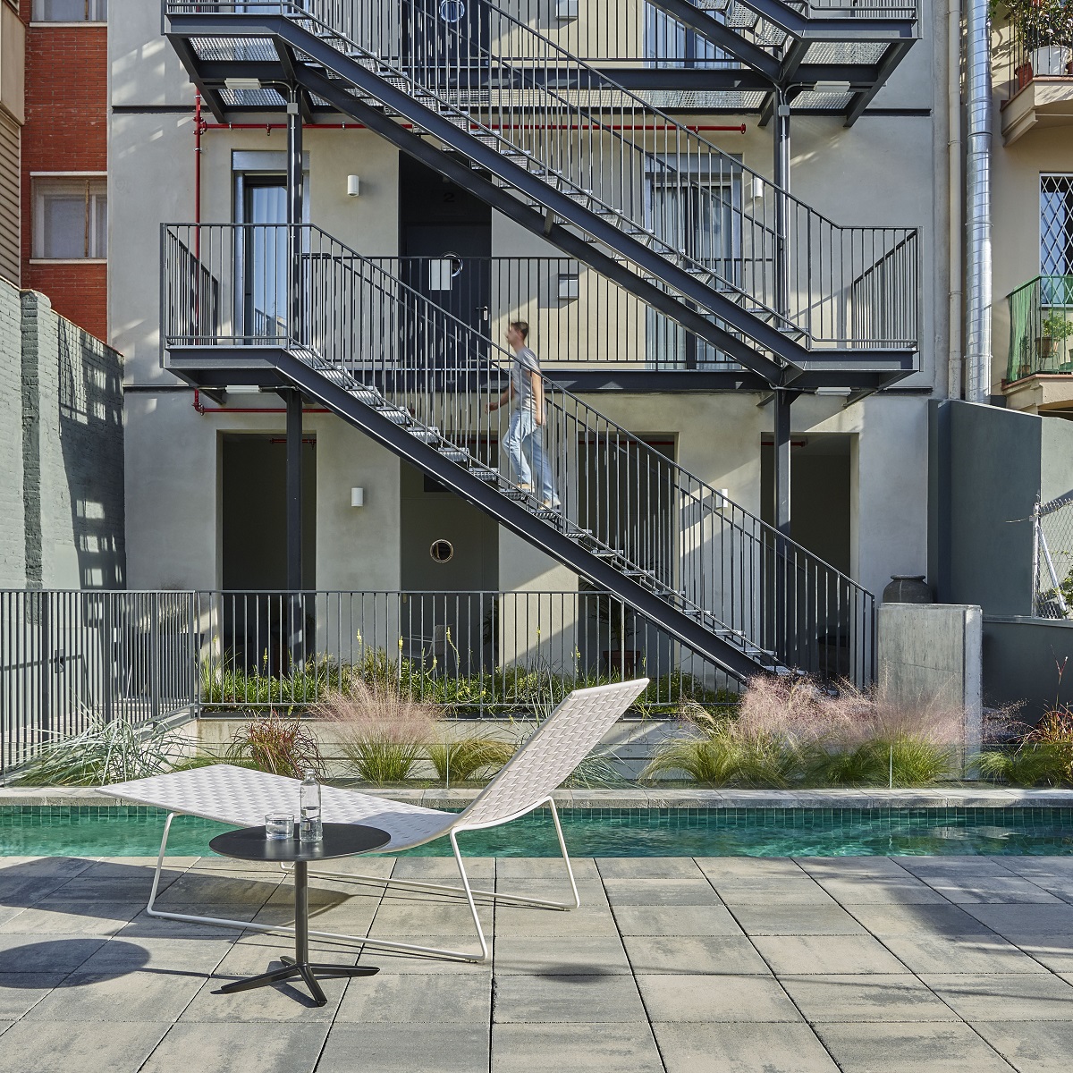 Andreu World Trenza Liege am Pool, moderne Outdoor Chaise Lounge mit Beistelltisch und Person auf Treppe.