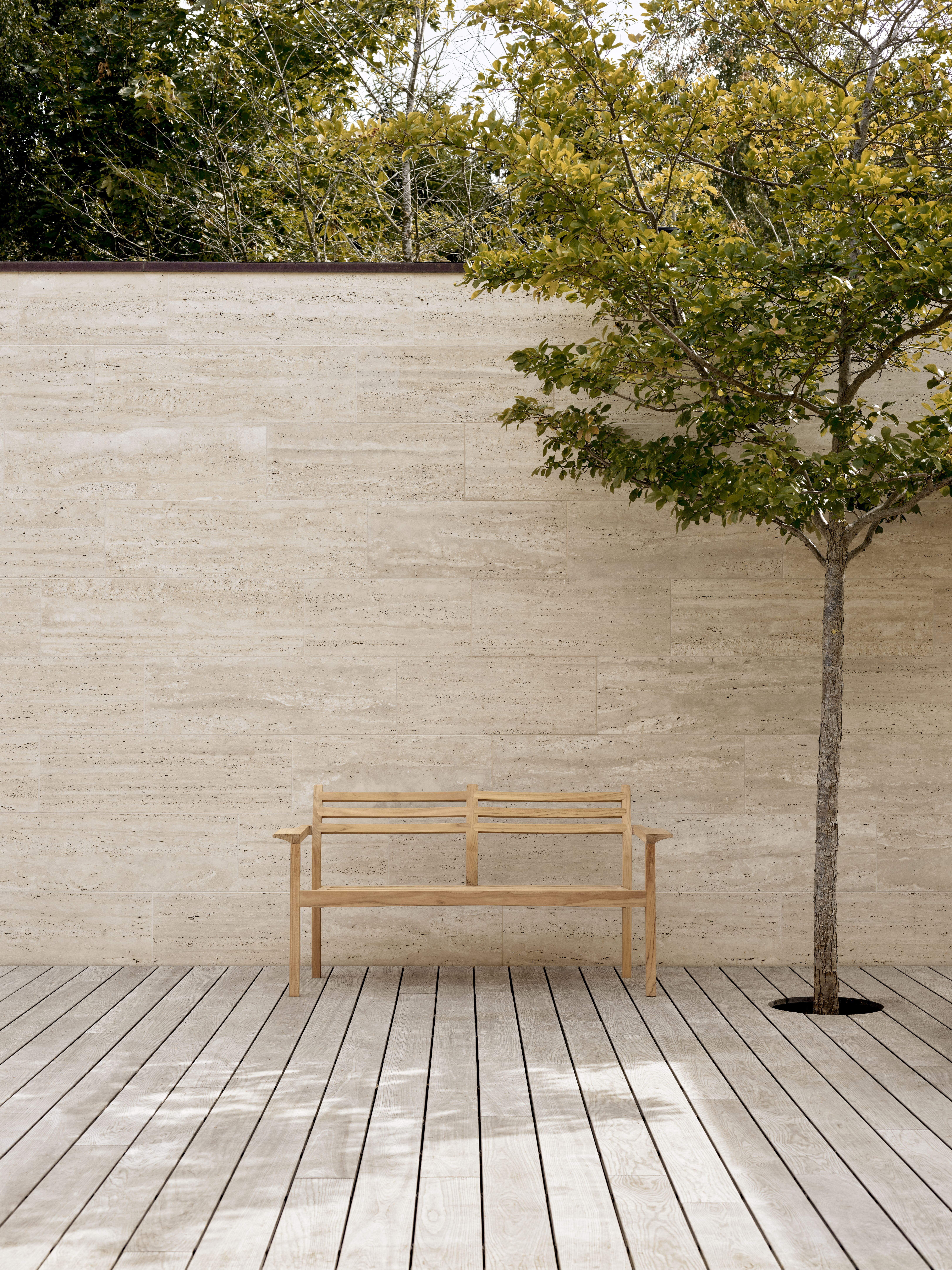 AH701 Carl Hansen Teak Gartenbank auf Holzterrasse vor Travertinwand und Baum.