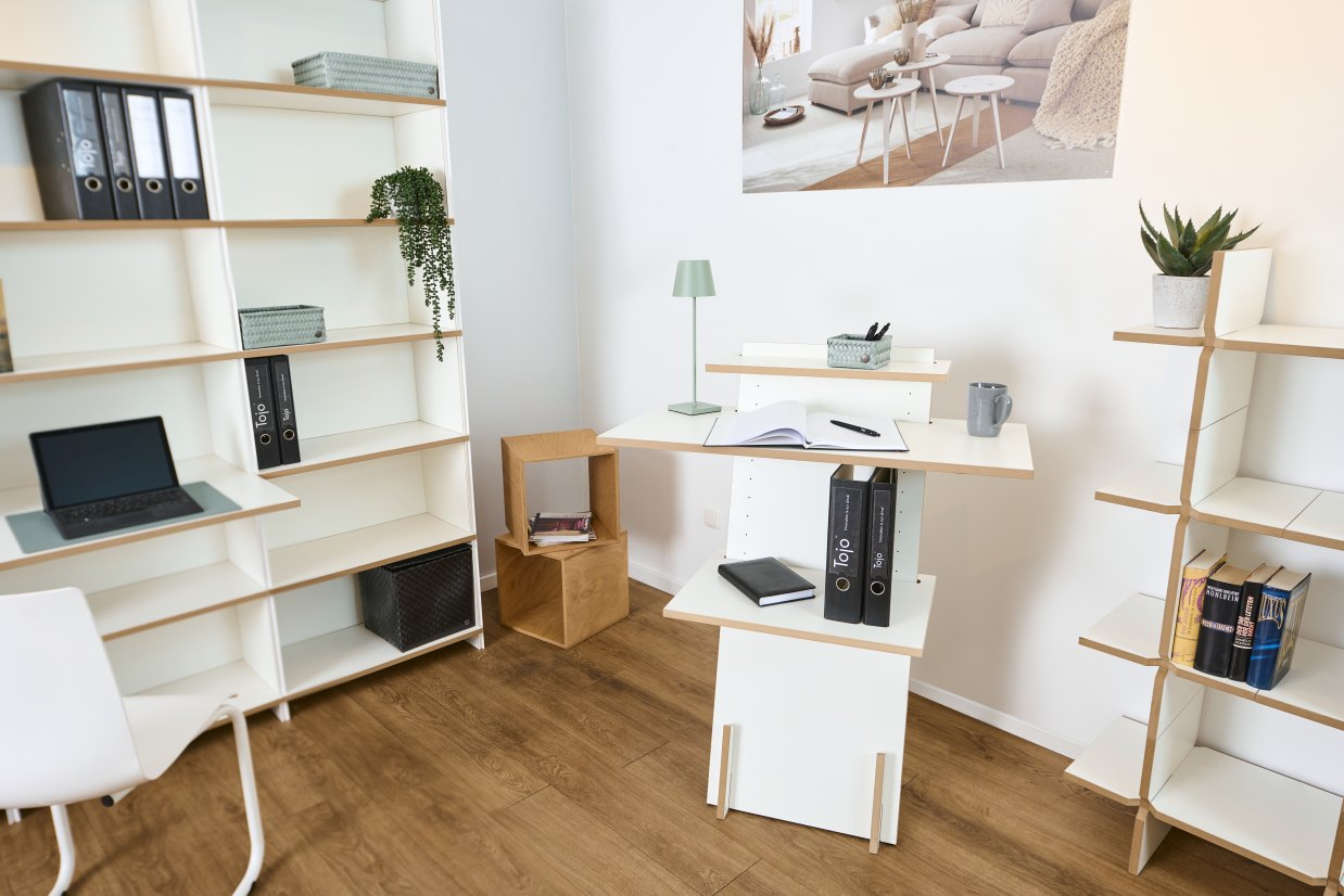 Helles Büro mit Tojo Stehpult aus Holz und weißen Regalen, Laptop, Ordnern und Dekoration.