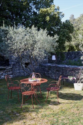 Fermob 1900 Gartentisch mit Stühlen in Rotbraun auf Rasen vor Steinmauer, Outdoor-Möbel für Garten.