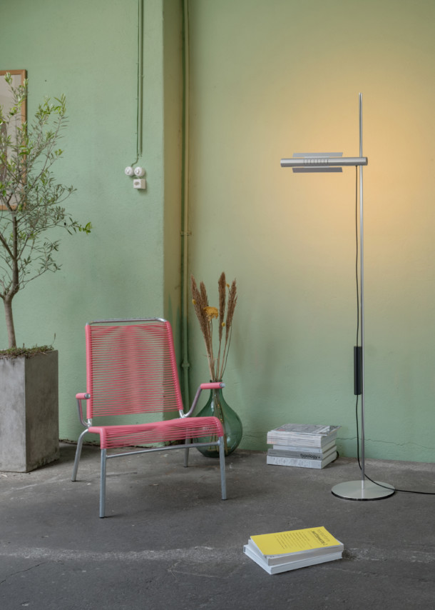 Moderne Stehlampe aus Metall neben rosa Outdoor-Stuhl und Bücherstapel vor mintgrüner Wand.