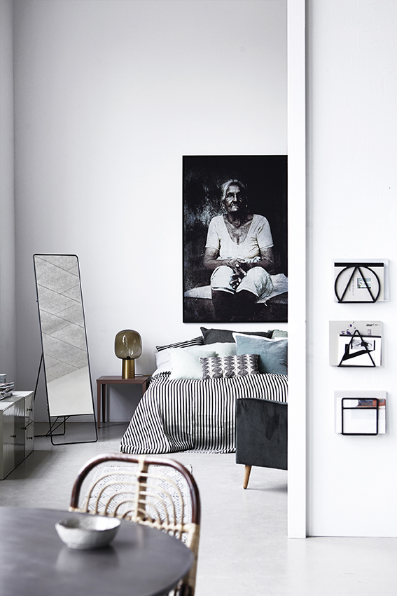 Helles Interieur mit Loge Zeitungsständer von House Doctor, Wanddeko, Spiegel und Bett mit gestreifter Tagesdecke.