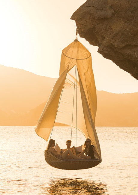 Drei Personen entspannen in einem hängenden Outdoor-Sessel mit Sonnensegel über dem Meer bei Sonnenuntergang.