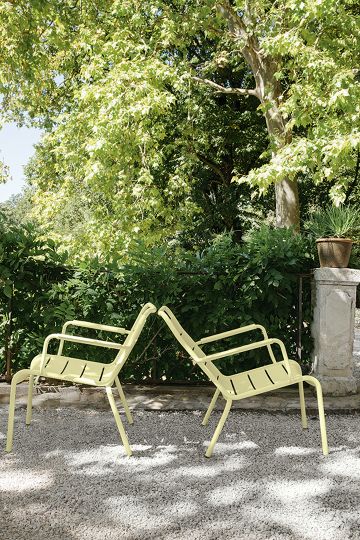 Zwei gelbe Fermob Luxembourg Lounge Sessel Outdoor auf Kies vor grüner Hecke und Baum.
