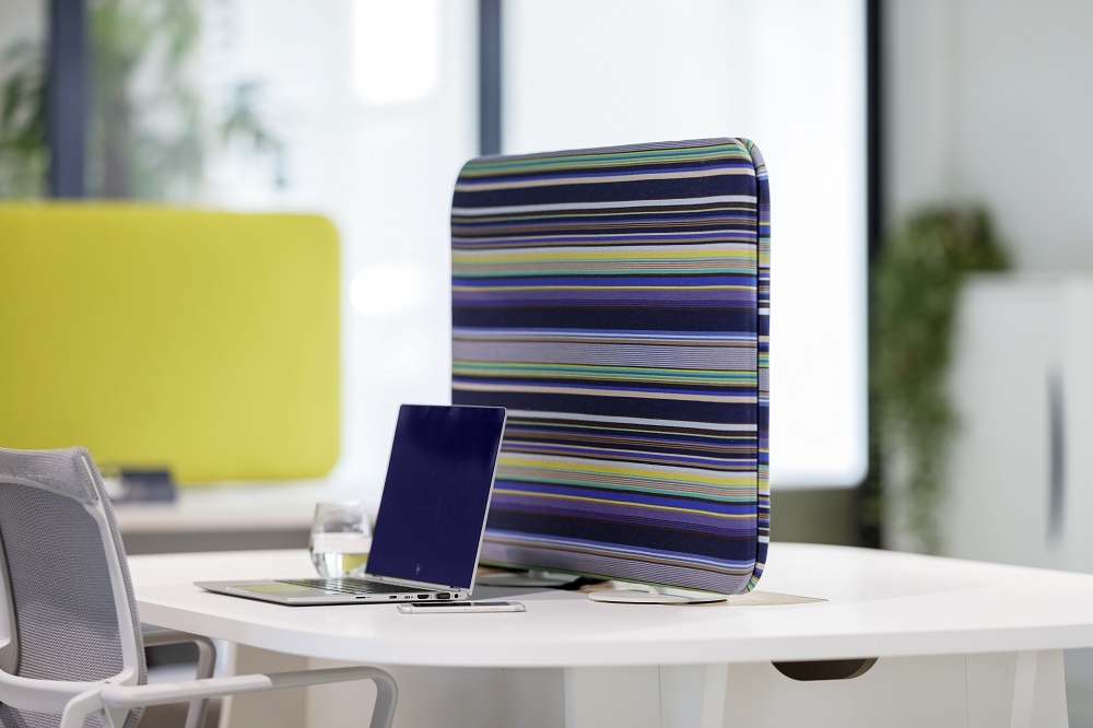 Cascando Pillow Desk Akustikelement mit bunten Streifen auf weißem Schreibtisch neben Laptop und Glas.