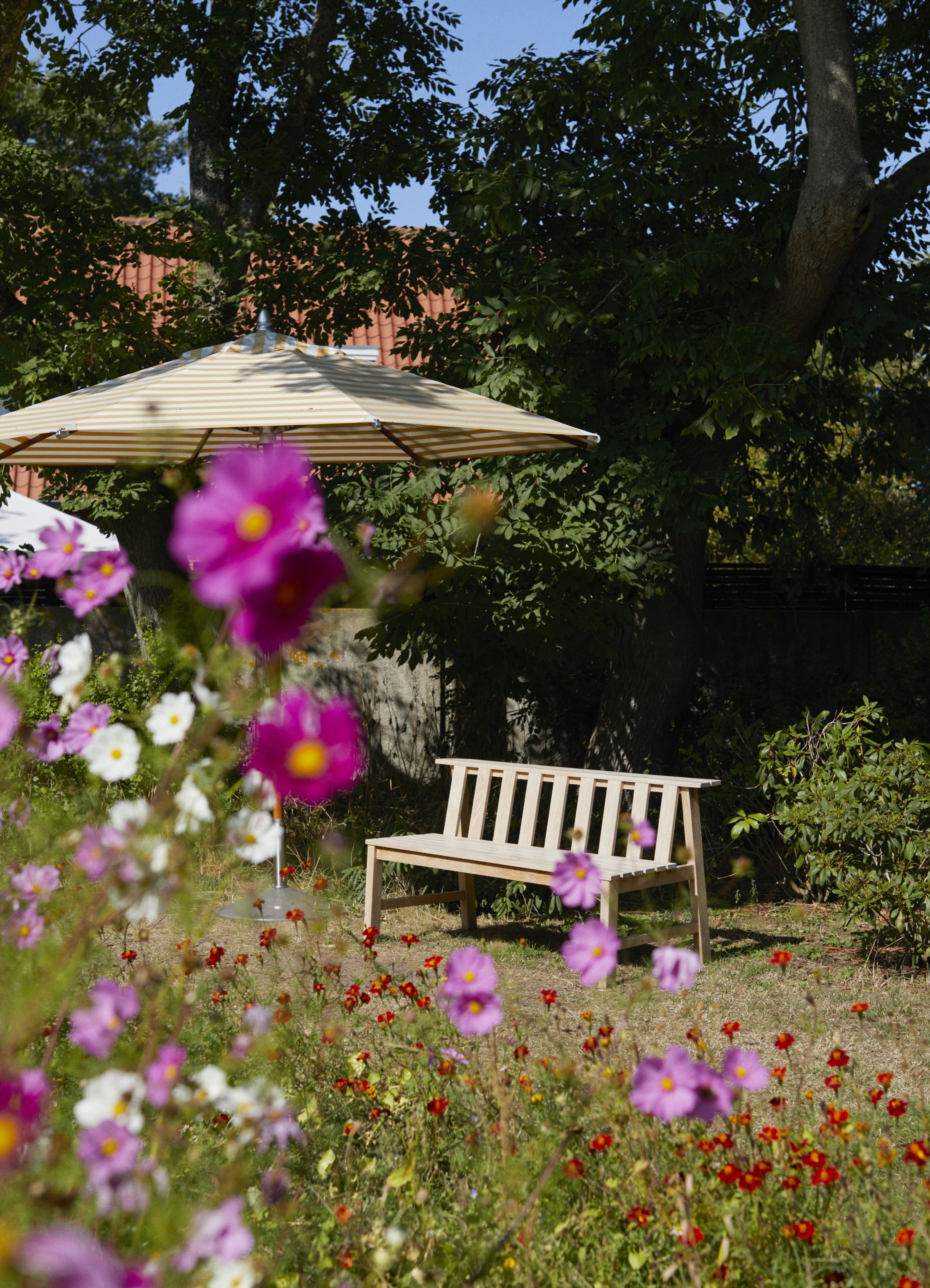 Cadence Teak Bank von Skagerak by Fritz Hansen im Garten mit Blumen und Sonnenschirm.