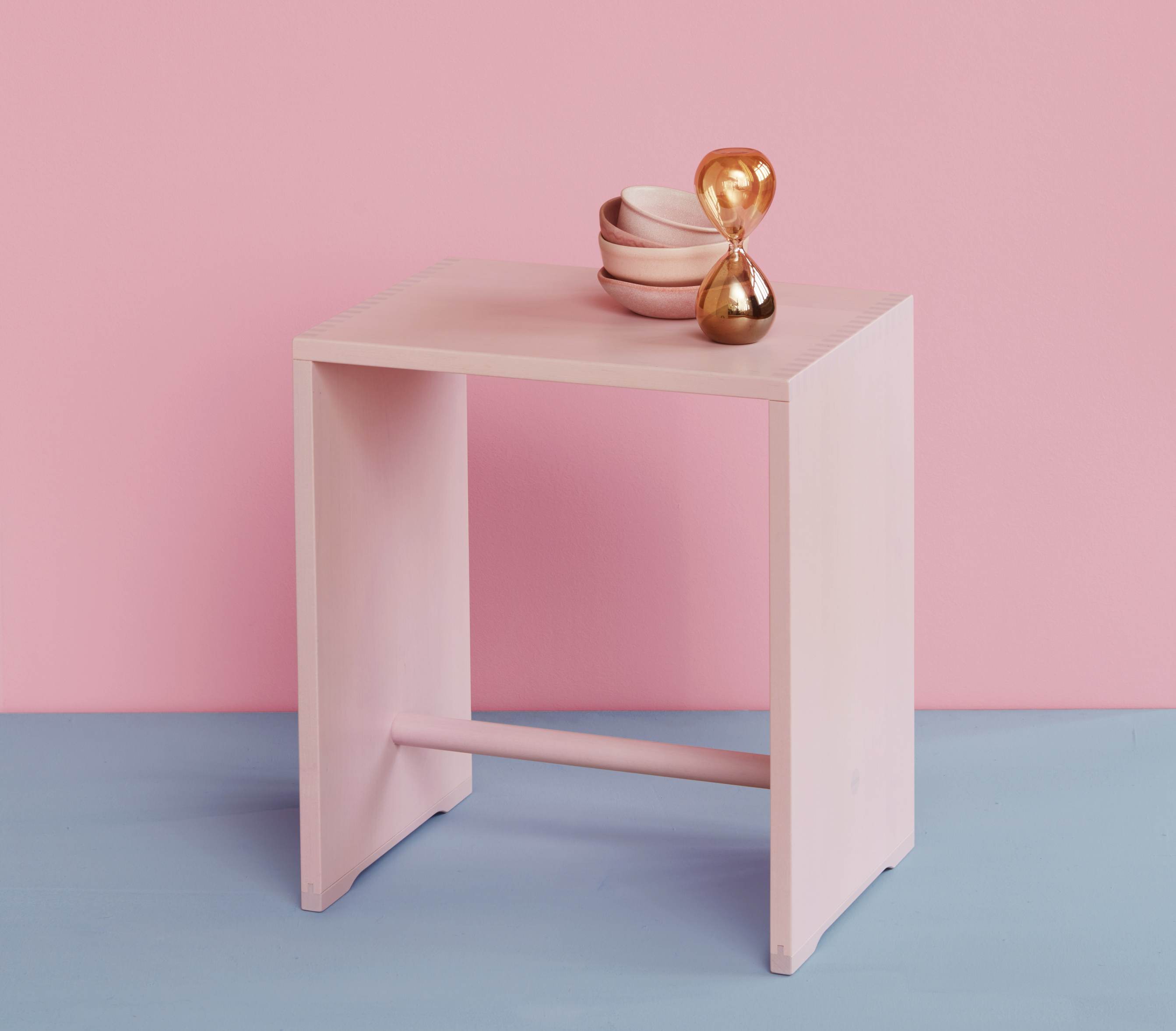 Rosa Hocker aus Holz mit Schalen und goldener Sanduhr vor rosa und blauem Hintergrund.