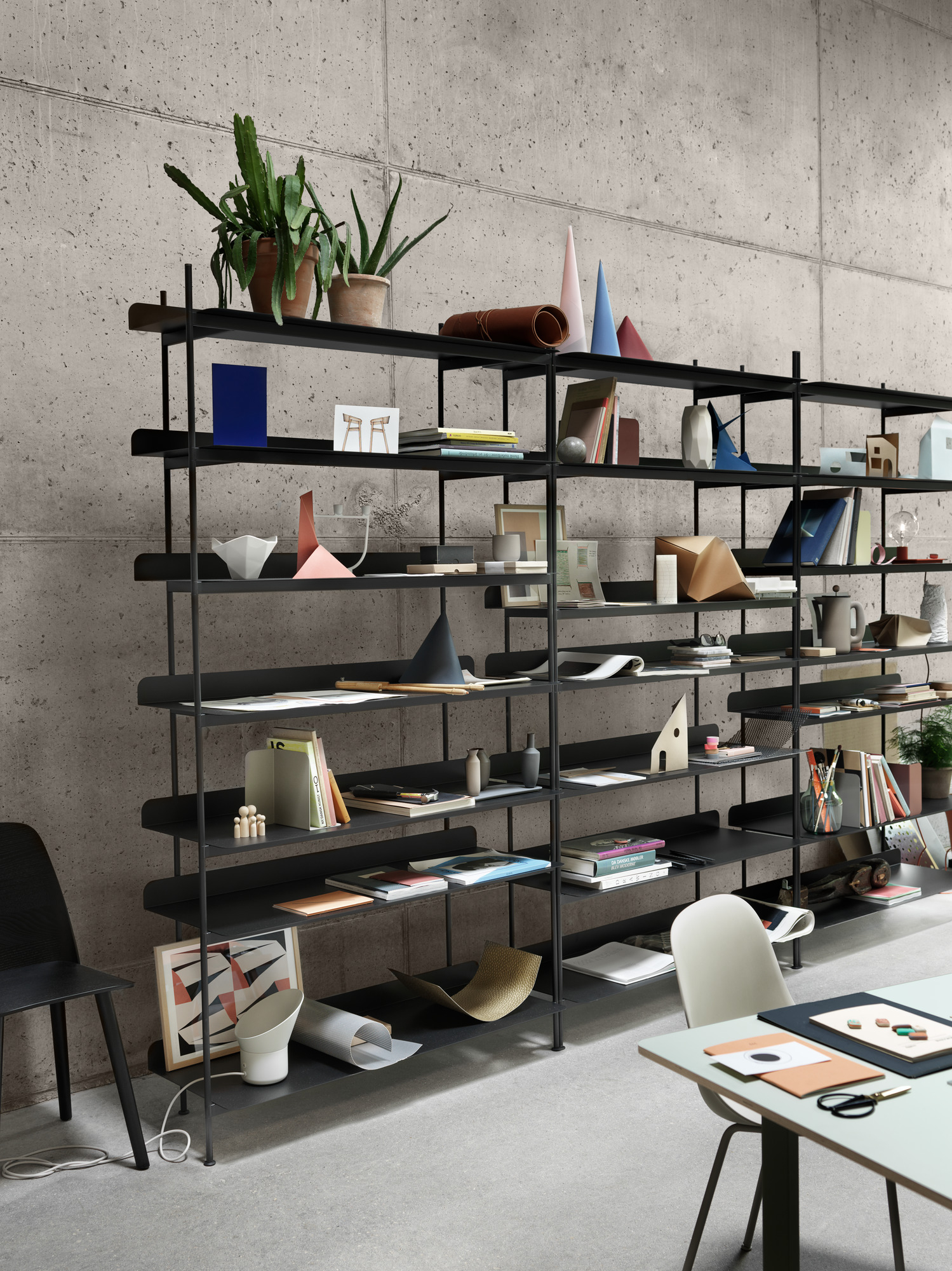 Schwarzes Regalsystem vor Betonwand, dekoriert mit Pflanzen, Büchern und Deko-Objekten, im Vordergrund ein 70/70 Tisch von Muuto.