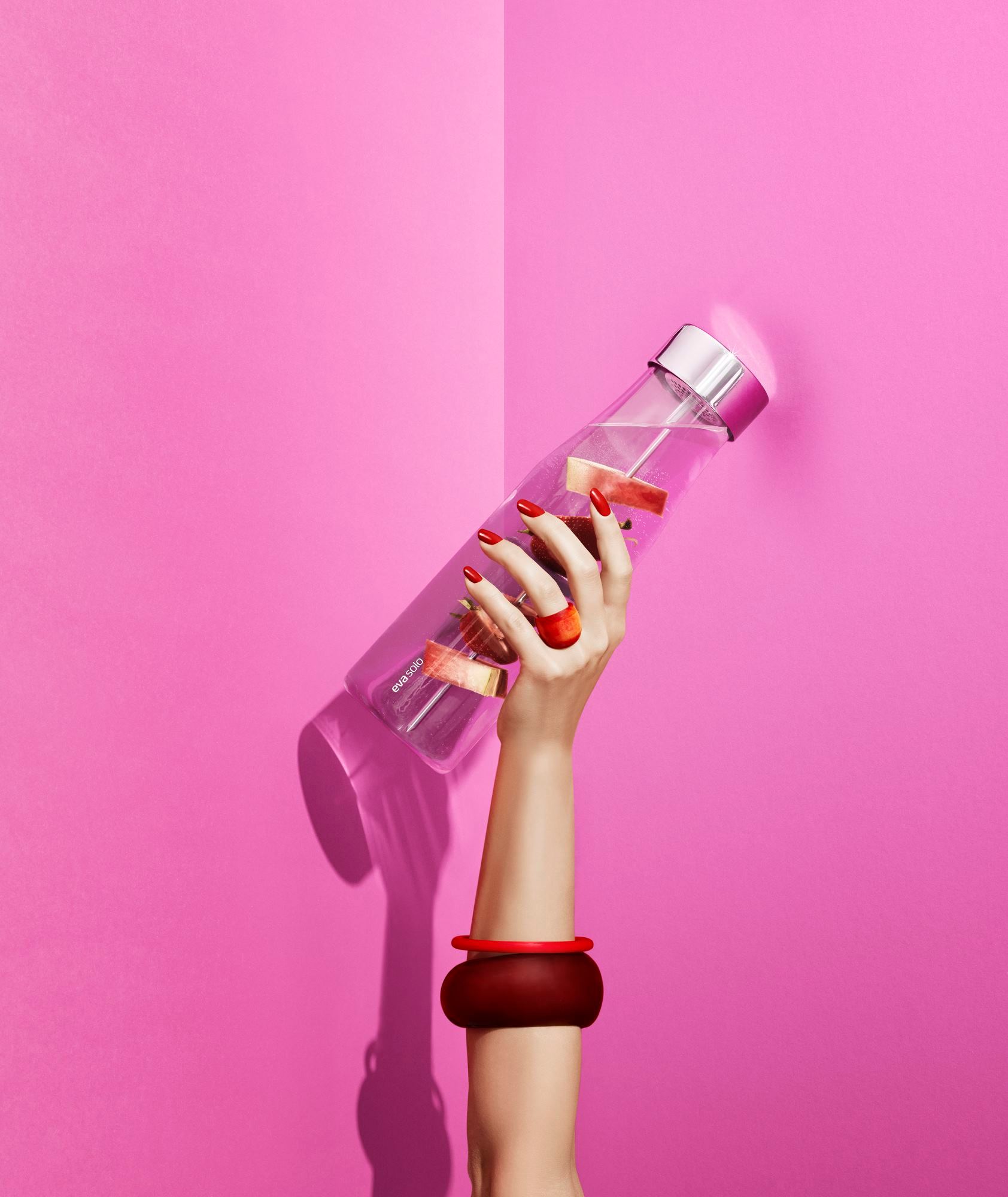 Frau hält Eva Solo My Flavour Karaffe mit Fruchtstücken vor pinkfarbenem Hintergrund. Stilvolle Wasserflasche für Infused Water.