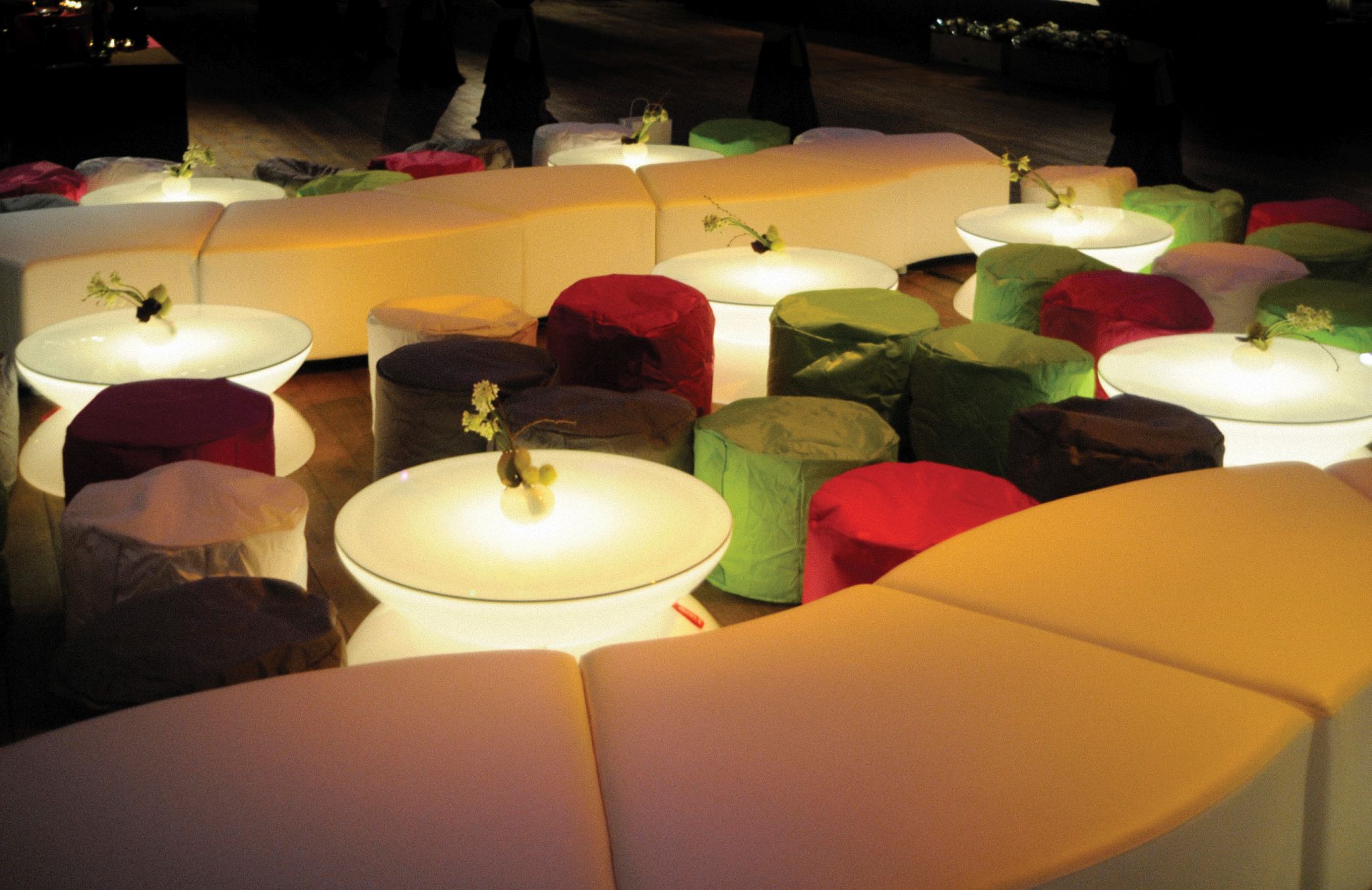 Innenaufnahme: Mehrere leuchtende Moree Lounge Tische mit bunten Sitzhockern und floraler Dekoration.