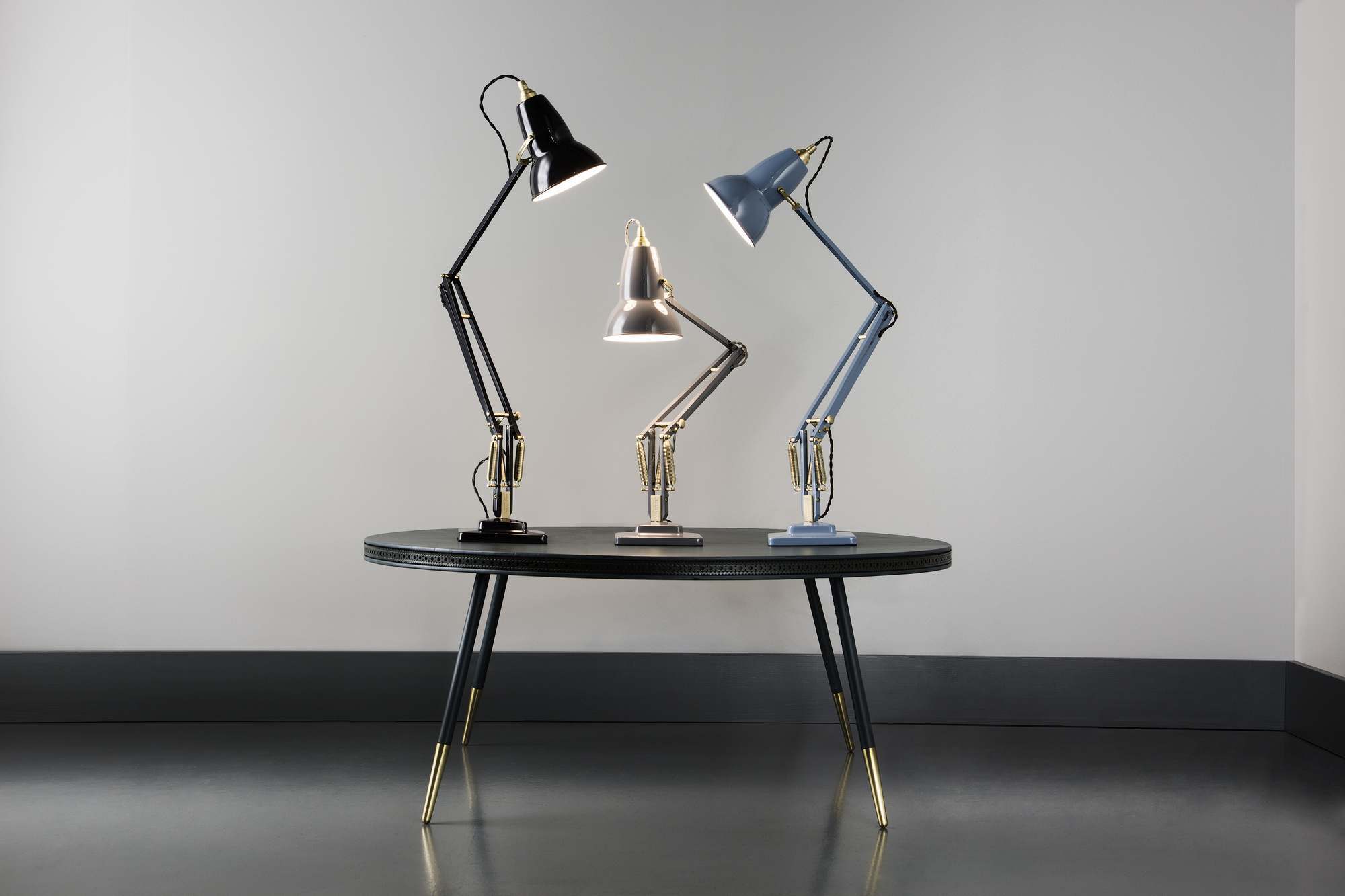 Drei Anglepoise Original 1227 Messing Tischleuchten in Schwarz, Silber und Blau auf einem Tisch.
