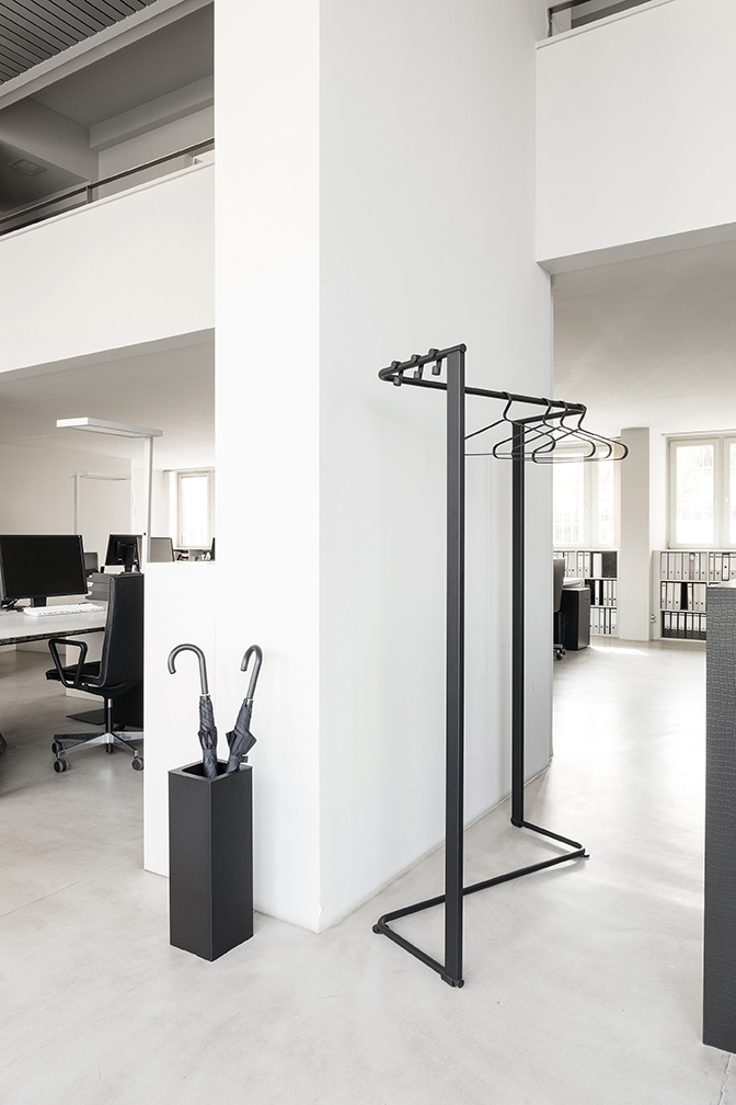 Schwarzer Schönbuch Kleiderbügel "Angle" im minimalistischen Design mit Kleiderbügeln und Schirmständer in modernem Büro.