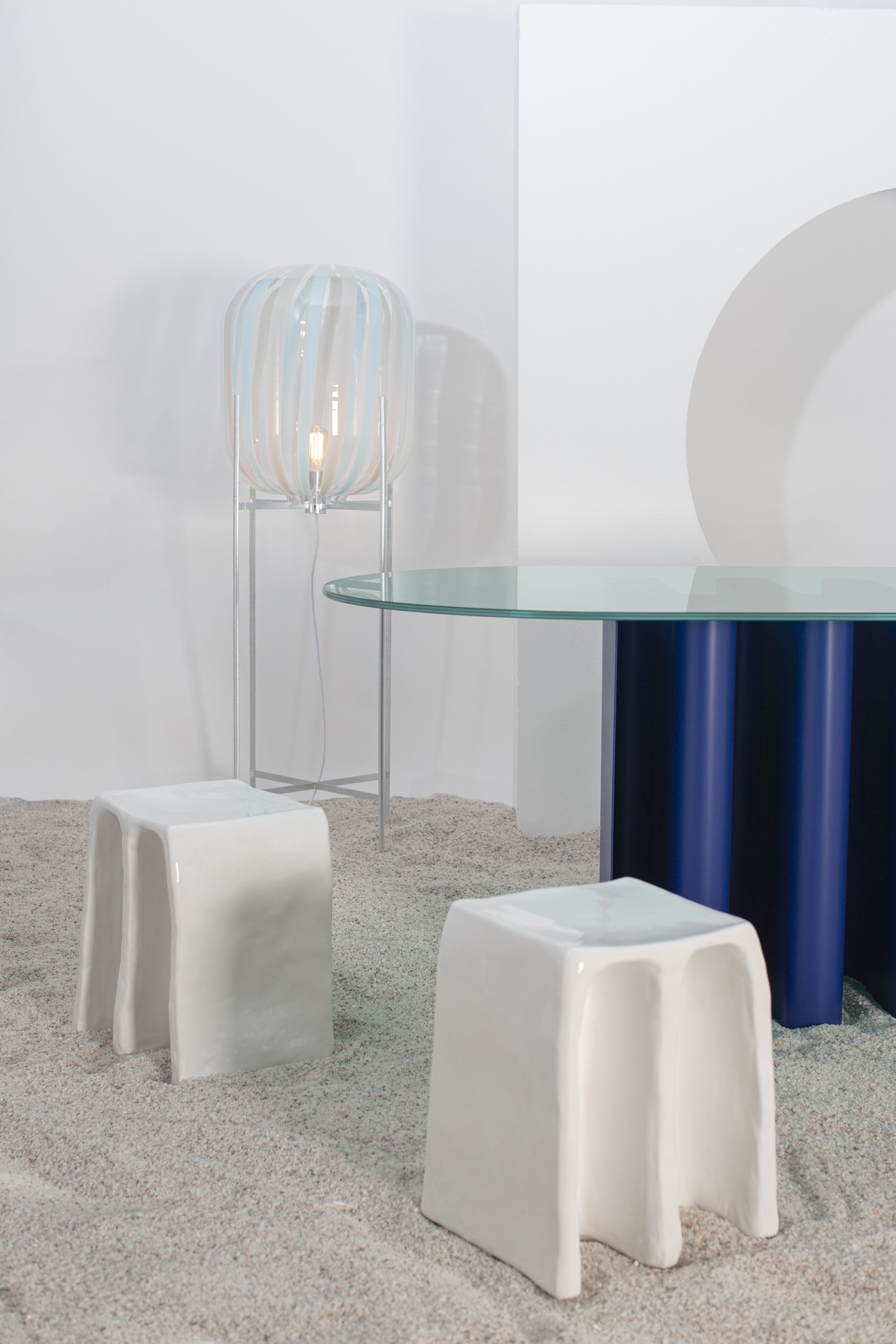 Tavolino2 Beistelltisch von Pulpo, zwei weiße Hocker und eine Stehlampe mit Glasfuß.