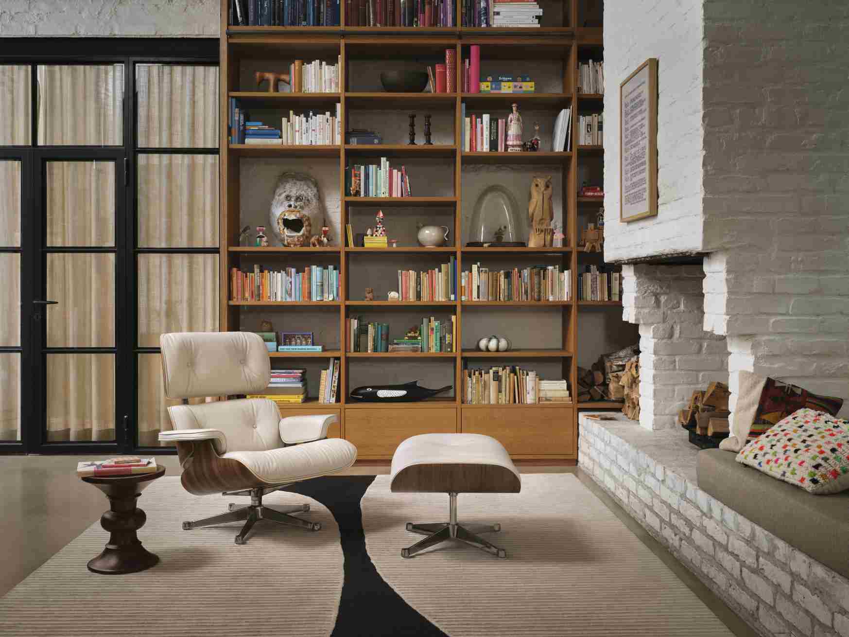 Eames Lounge Chair mit Hocker in hellem Leder vor Bücherregal und Kamin.