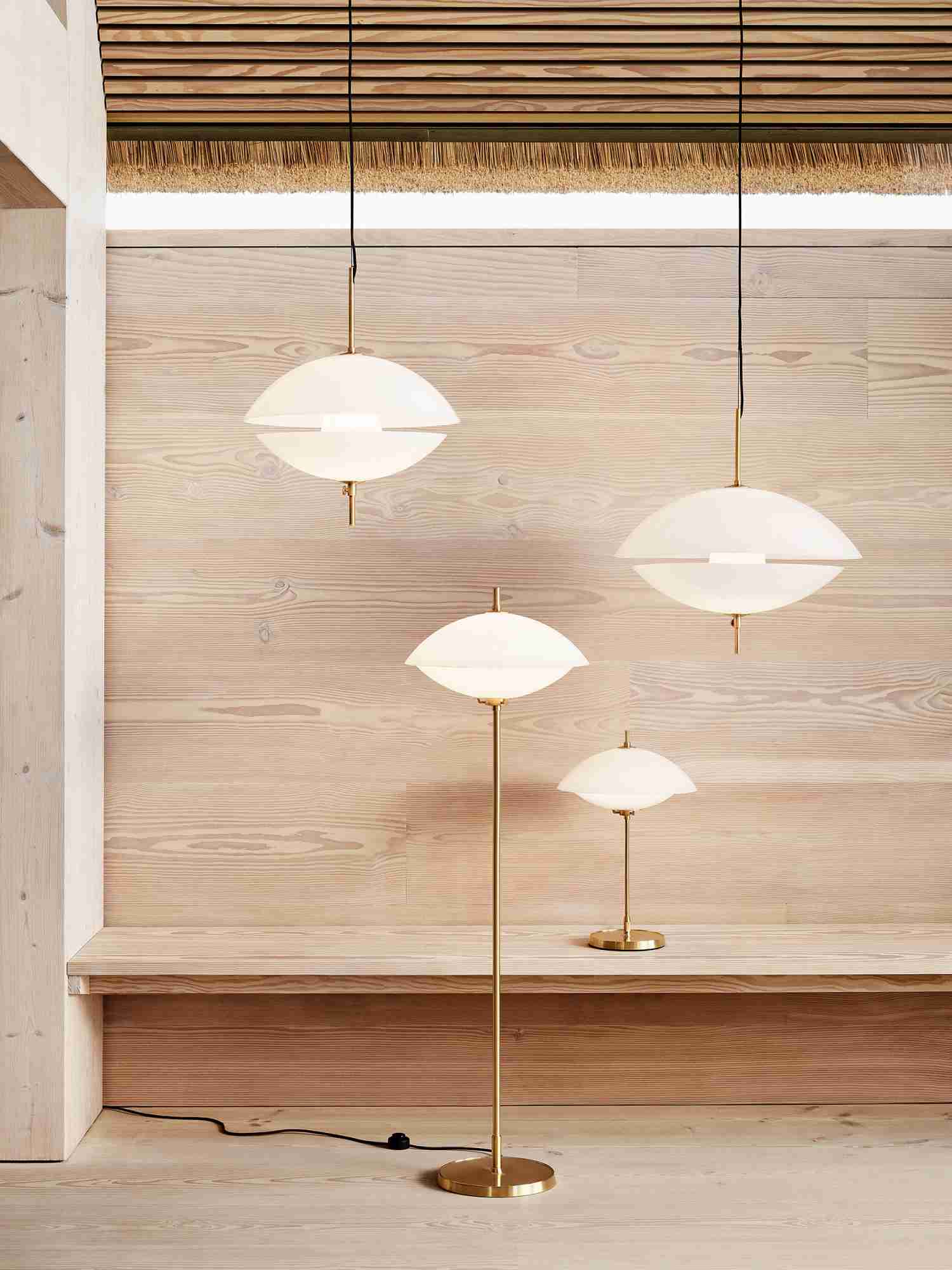 Nahaufnahme der Clam Tischleuchte von Fritz Hansen, Stehleuchte und Pendelleuchten mit Messingdetails vor Holzwand.